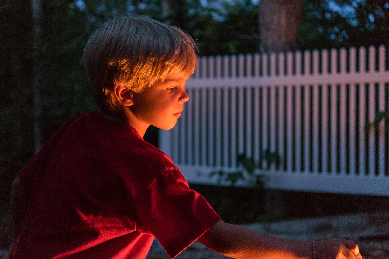 A child in a red shirt roasts marshmallows over a fire, focused on making s'mores at dusk.