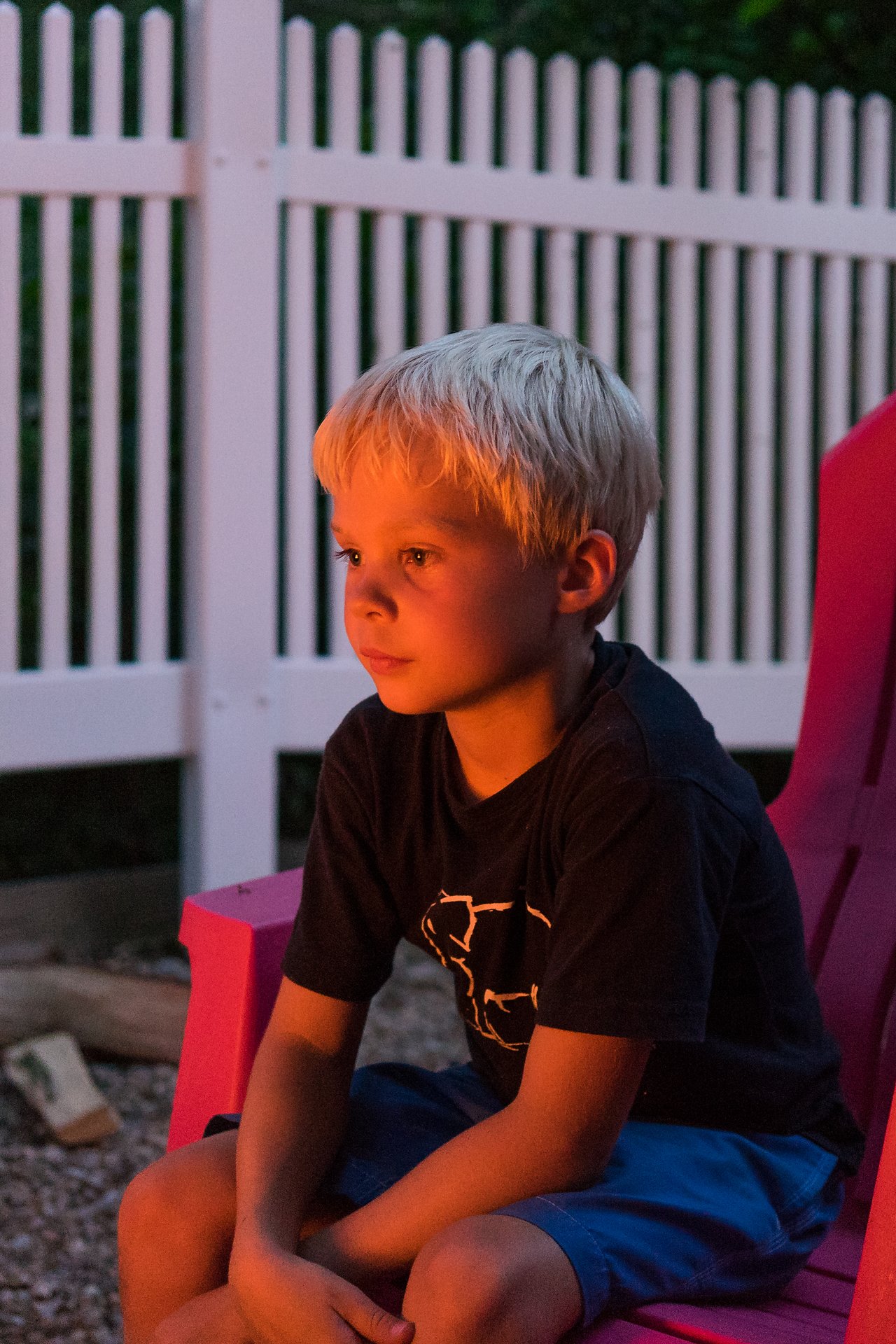 A young boy sits in a red chair, gazing at a fire, illuminated by its warm glow.