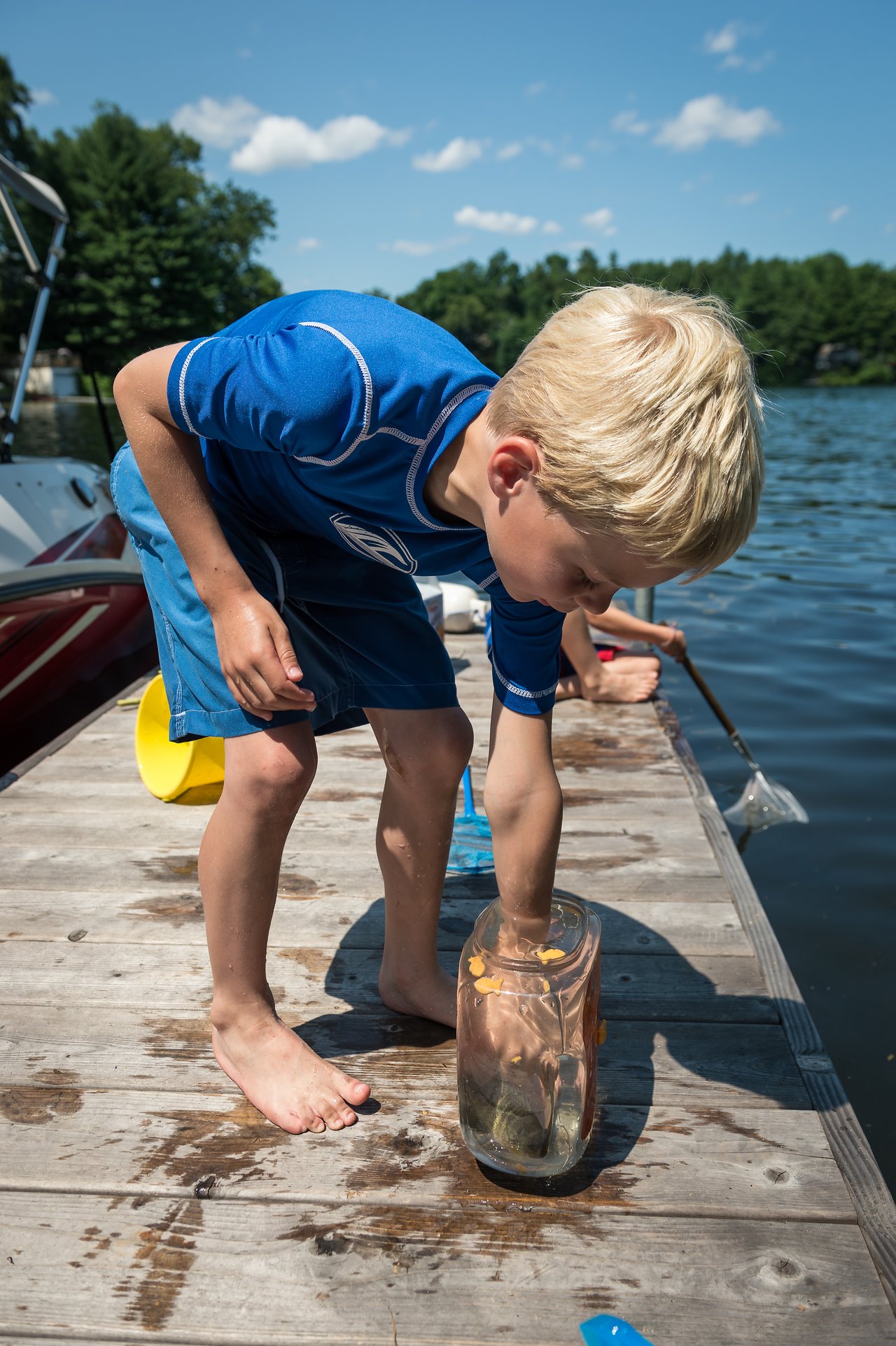 A young boy on a dock reaches into a jar of water, examining something inside.