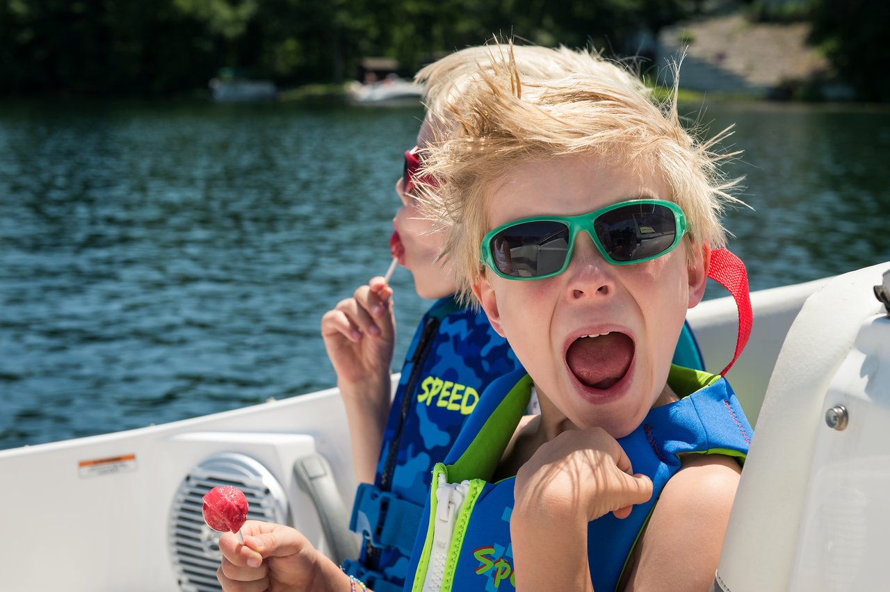 Two children wearing life jackets sit on a boat, one excitedly shouting while holding a lollipop.