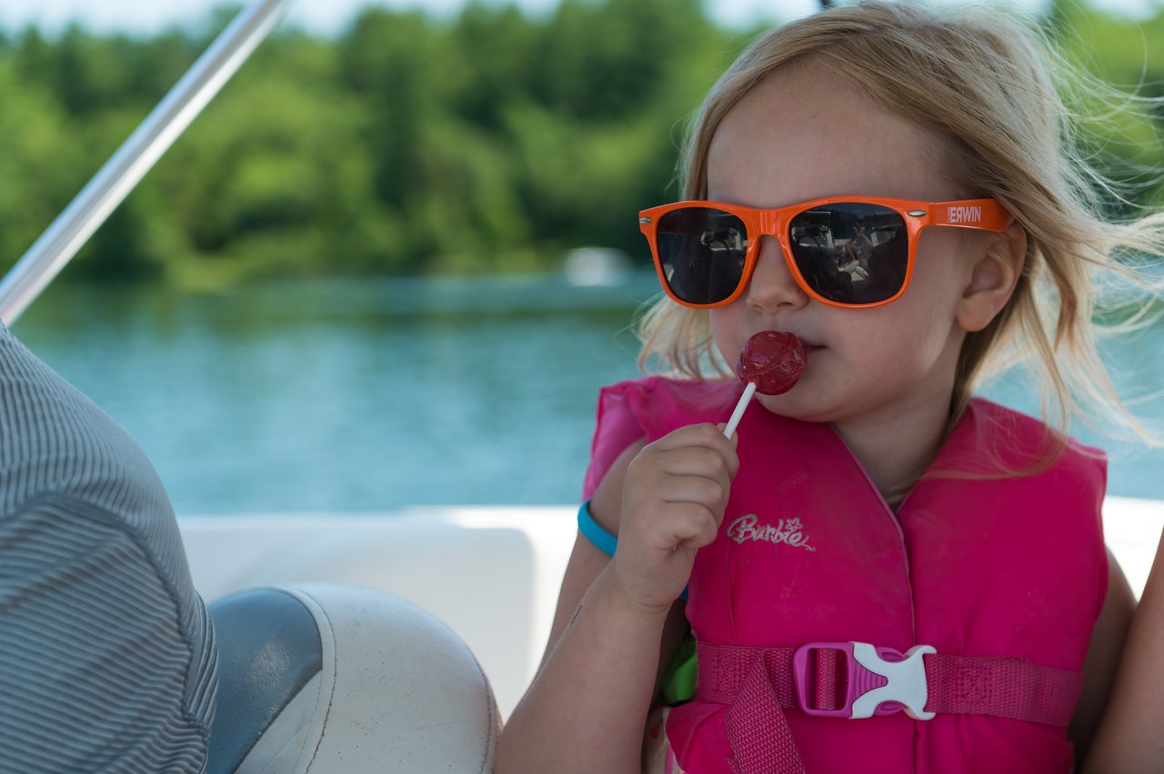 A young child wearing sunglasses and a life jacket enjoys a lollipop while sitting on a boat.