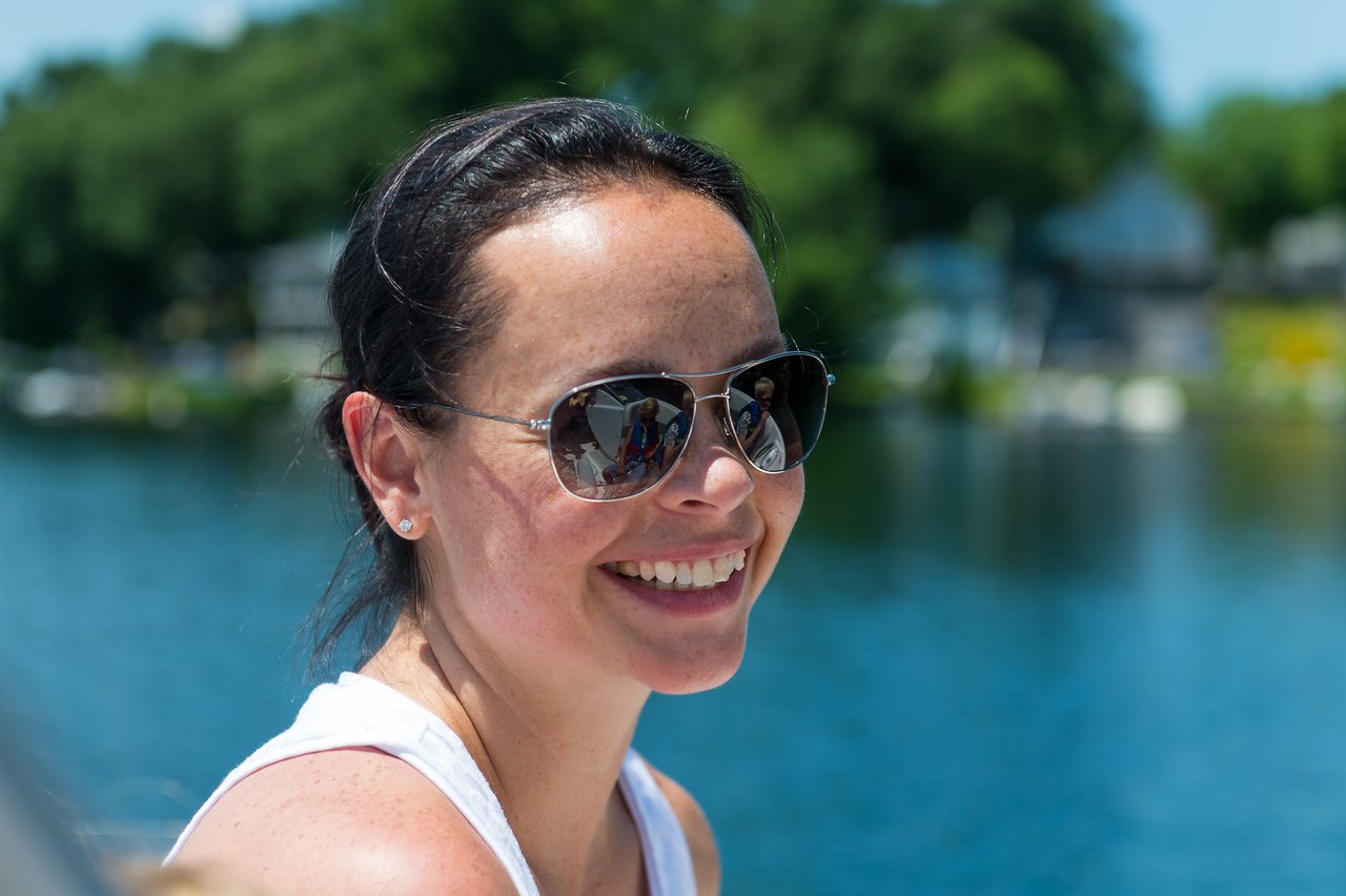 A woman wearing sunglasses and a white tank top smiles while sitting near the water on a sunny day.