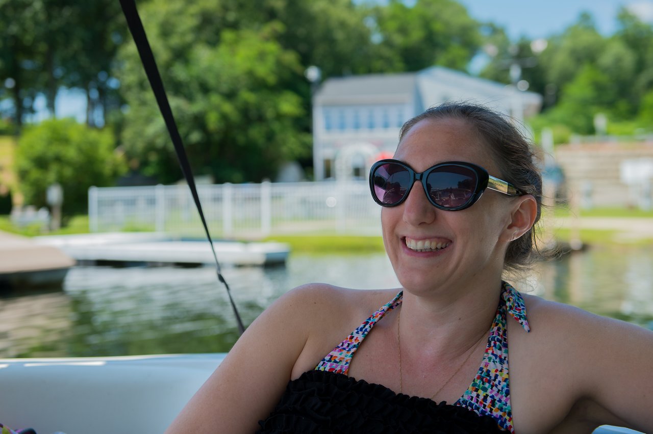 A woman wearing sunglasses and a swimsuit smiles while relaxing on a boat near the water.