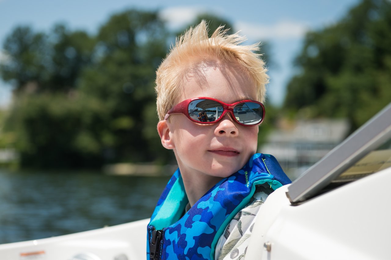 A child wearing red sunglasses and a blue life jacket sits on a boat, looking off to the side.