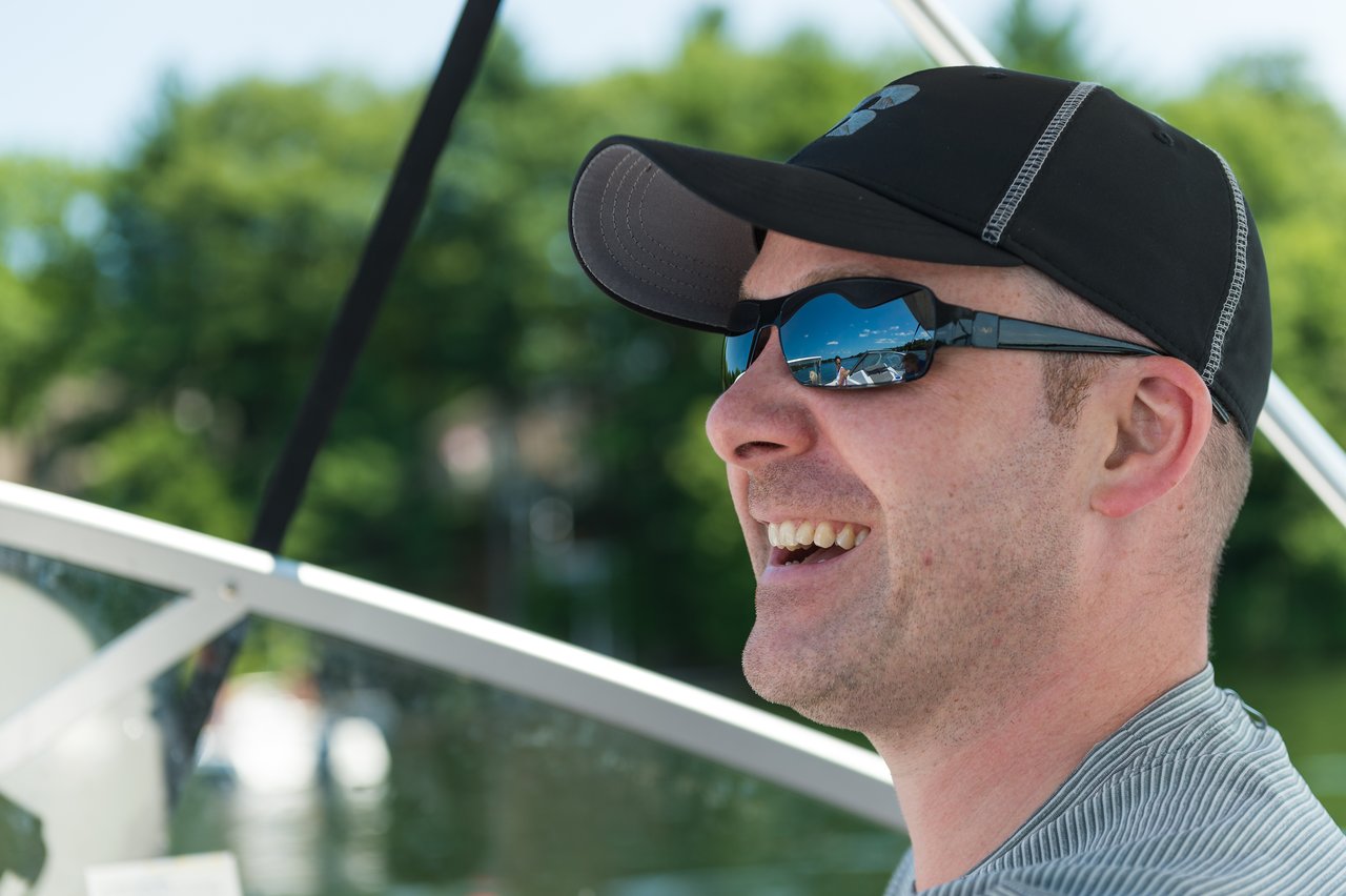 A man wearing sunglasses and a black cap smiles while riding on a boat.