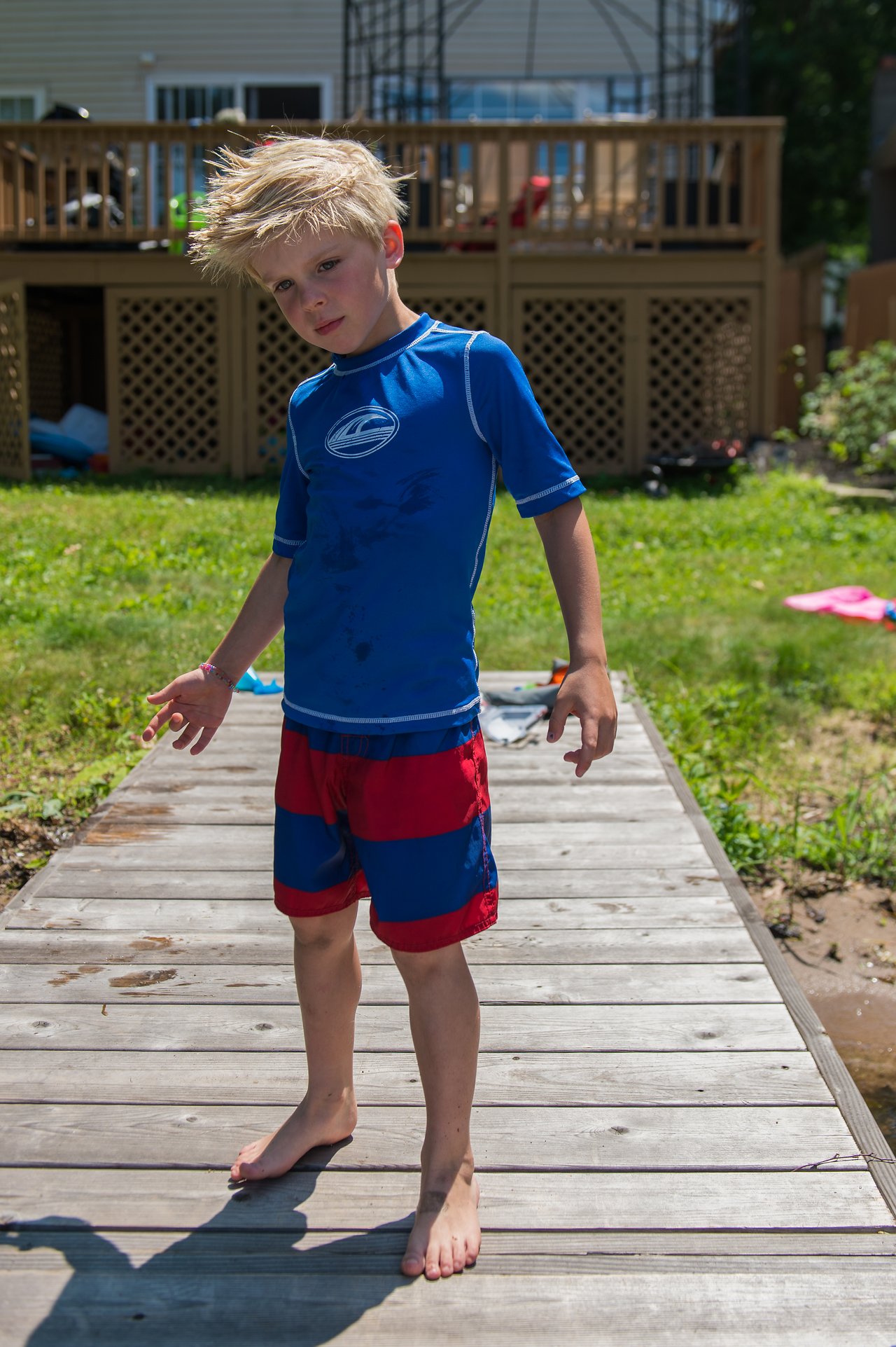 A young boy in a blue rash guard and red swim shorts stands barefoot on a wooden dock, slightly leaning.