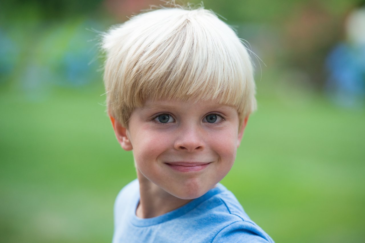 A young child with blonde hair and a blue shirt smiles at the camera in an outdoor setting.