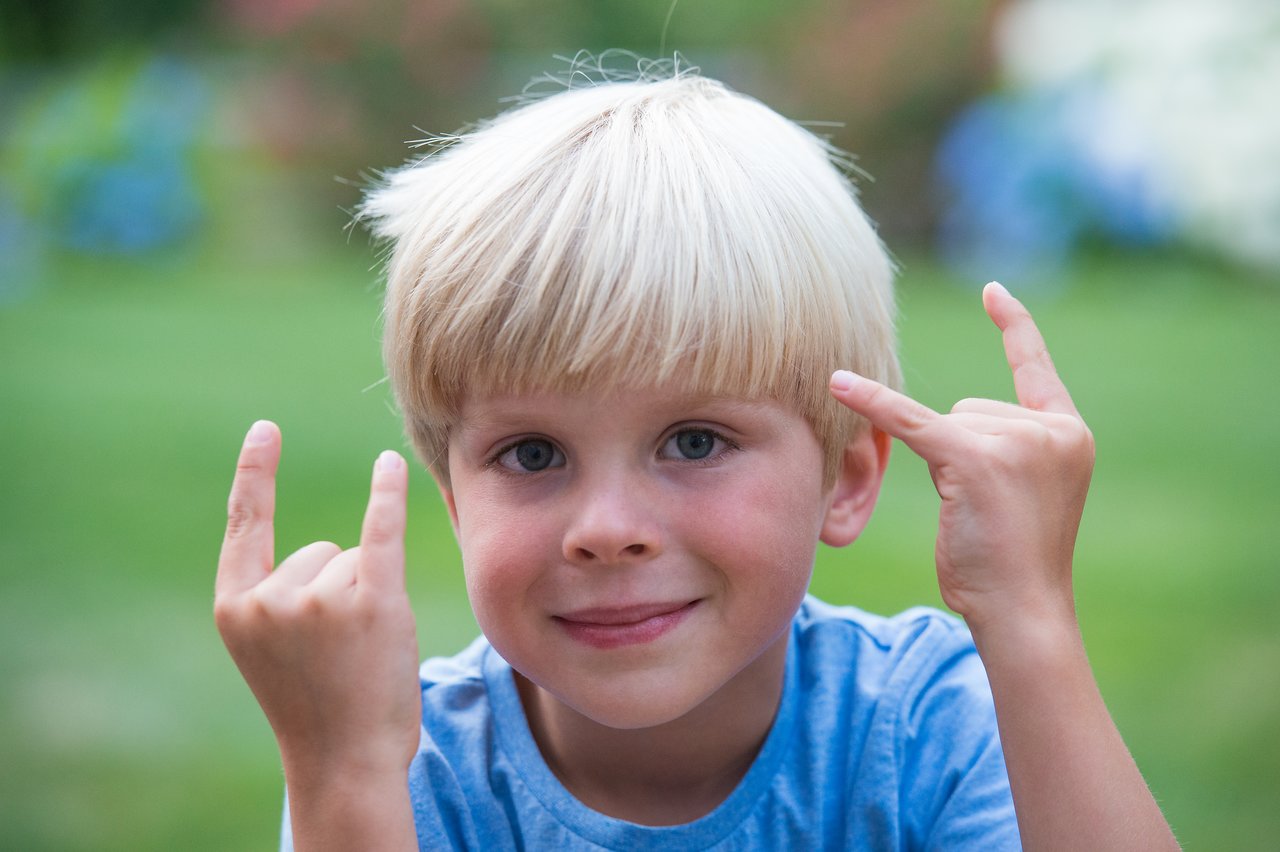 A young boy with blonde hair and a blue shirt smiles while making a hand gesture with both hands.