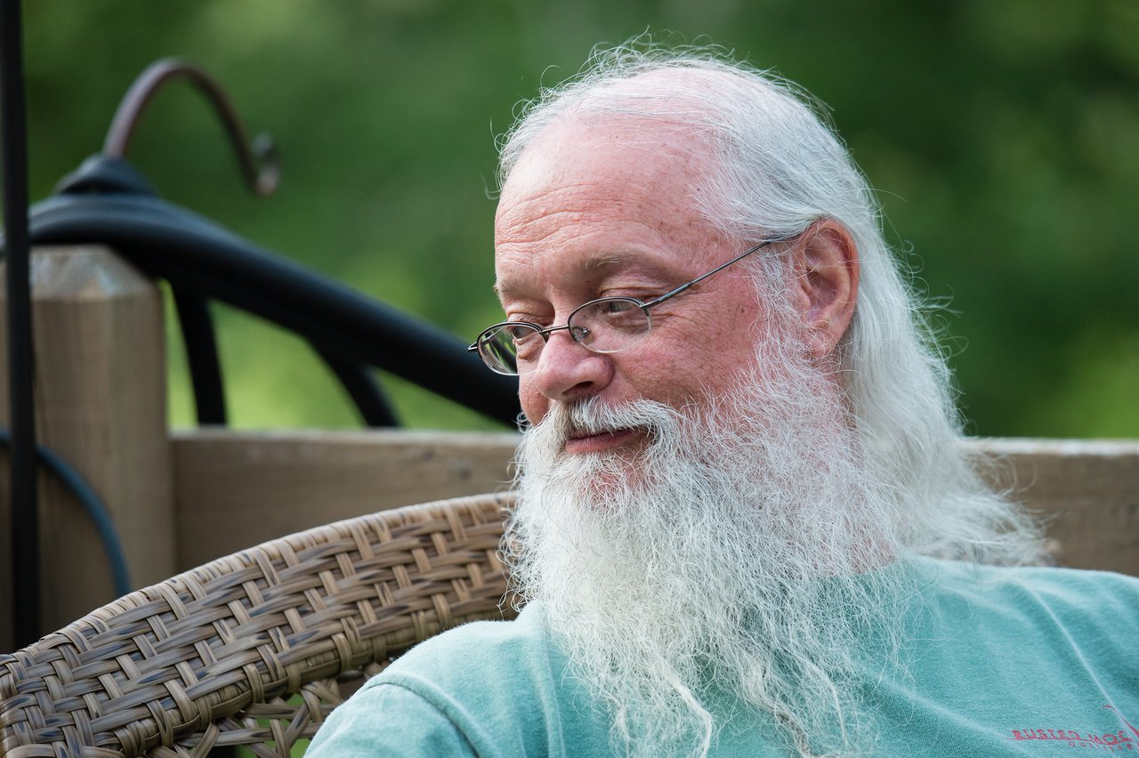 An older man with a long white beard and glasses sits outdoors, looking to the side with a slight smile.