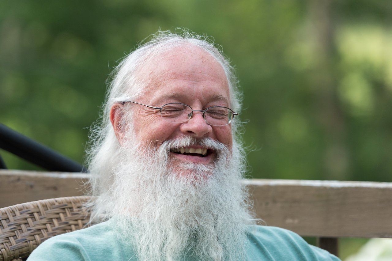 A man with a long white beard and glasses is sitting outdoors, smiling with his eyes closed.