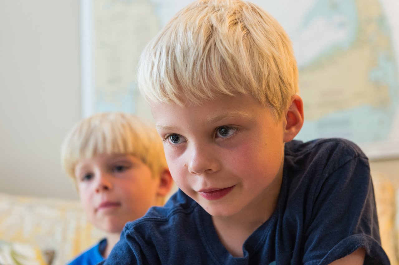 A boy in a blue shirt focuses intently while playing a game, with another child watching behind him.