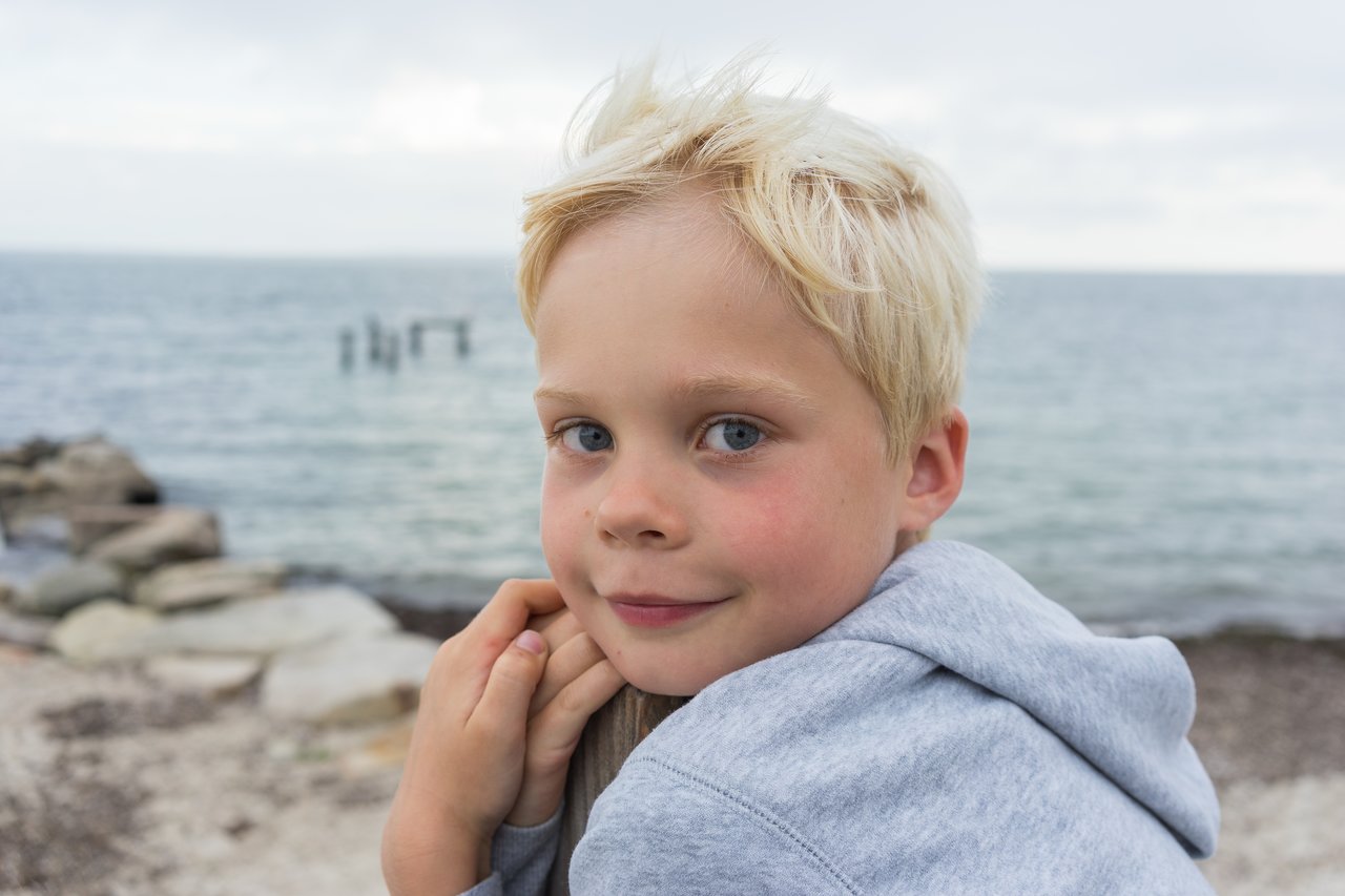 A young child in a gray hoodie leans on a wooden post, smiling near the beach with the ocean behind them.