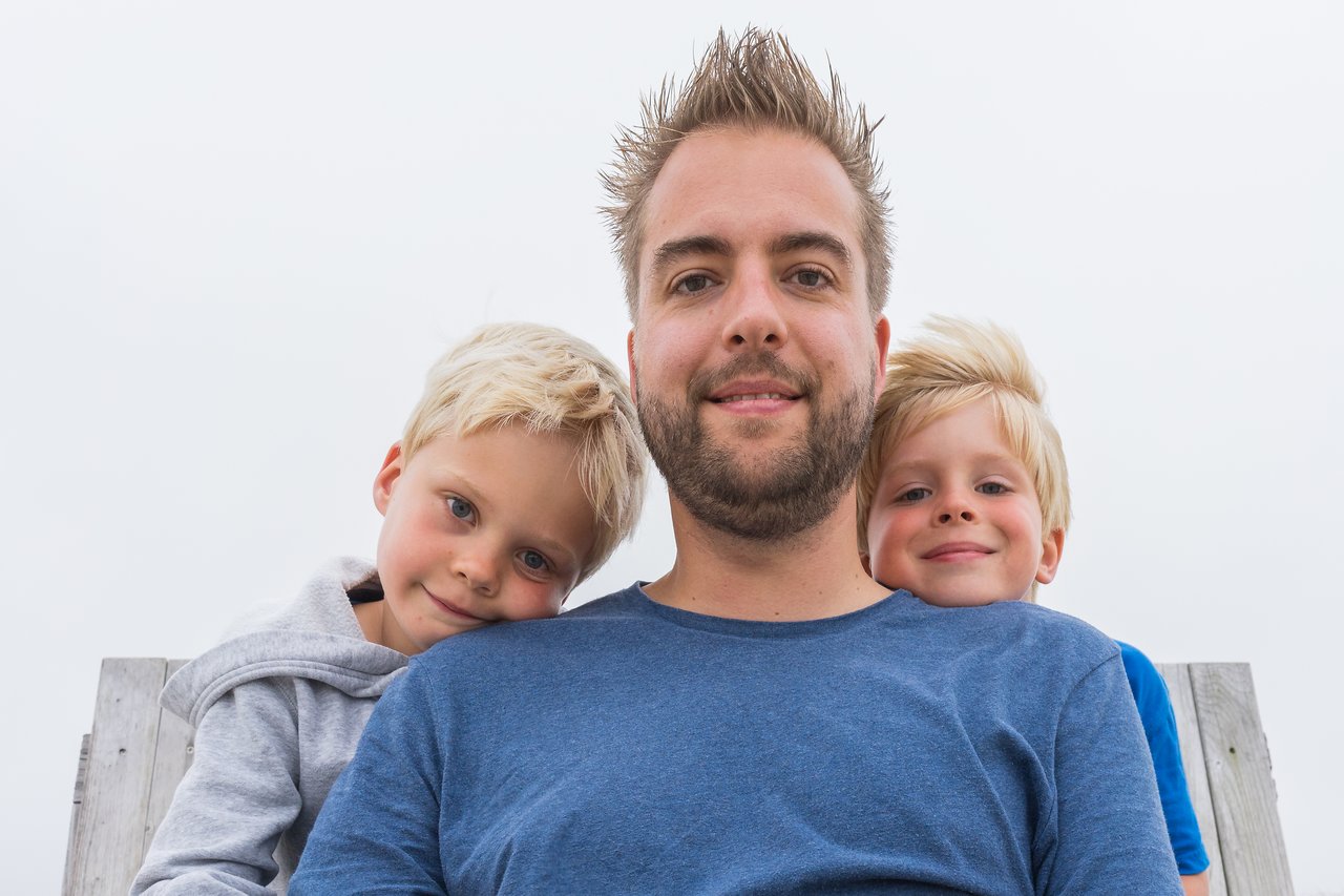 A man sits on a bench with two young children leaning on his shoulders, all smiling at the camera.