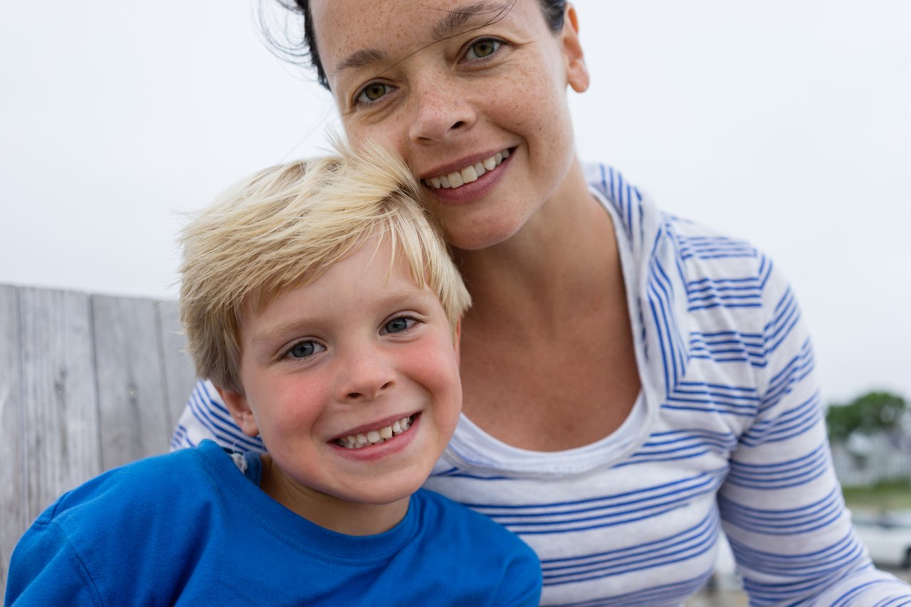 A woman and a child smile at the camera while sitting outdoors near a wooden fence.