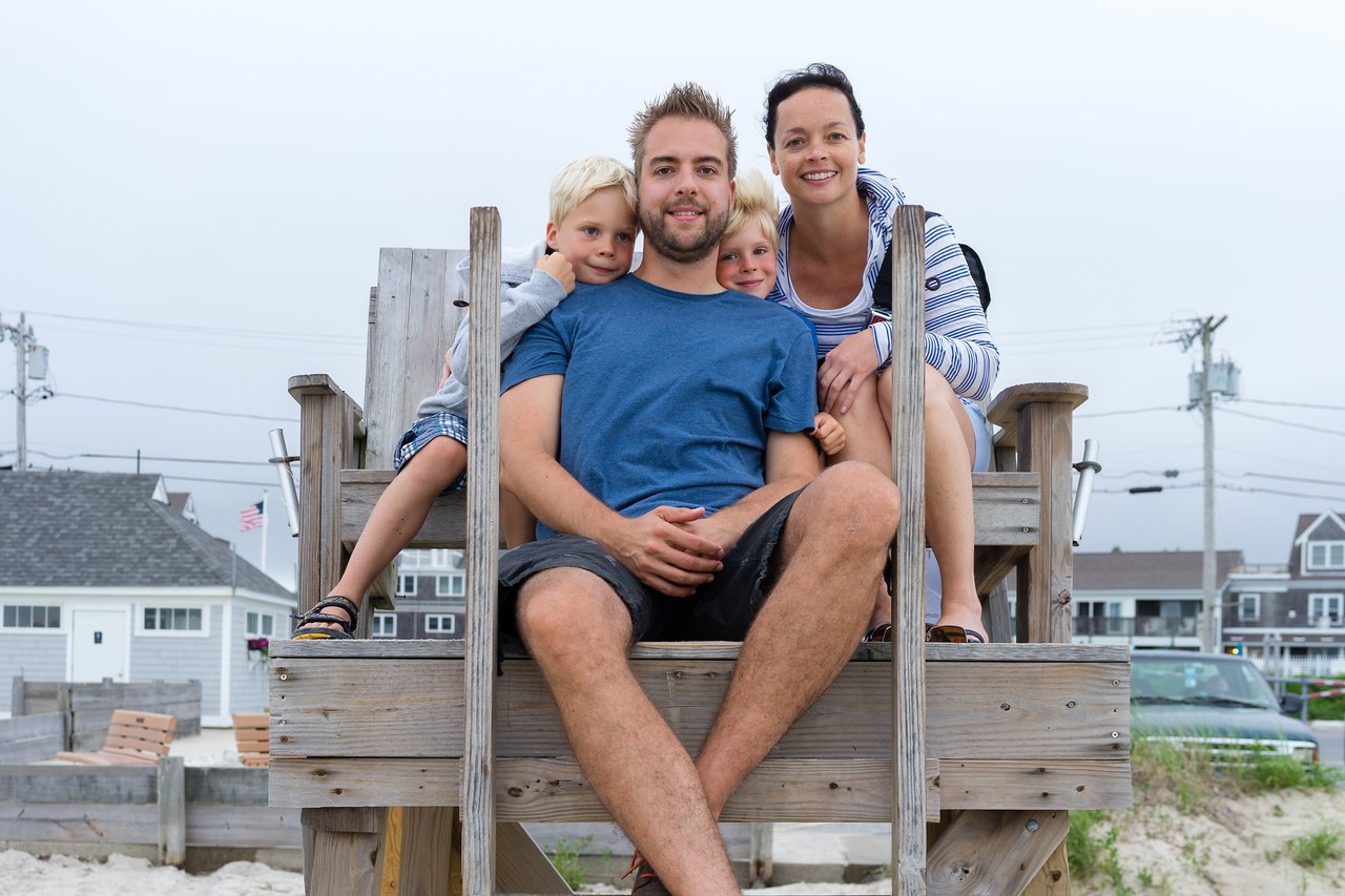 A family of four sits together on a wooden lifeguard chair, smiling at the camera on a cloudy beach day.