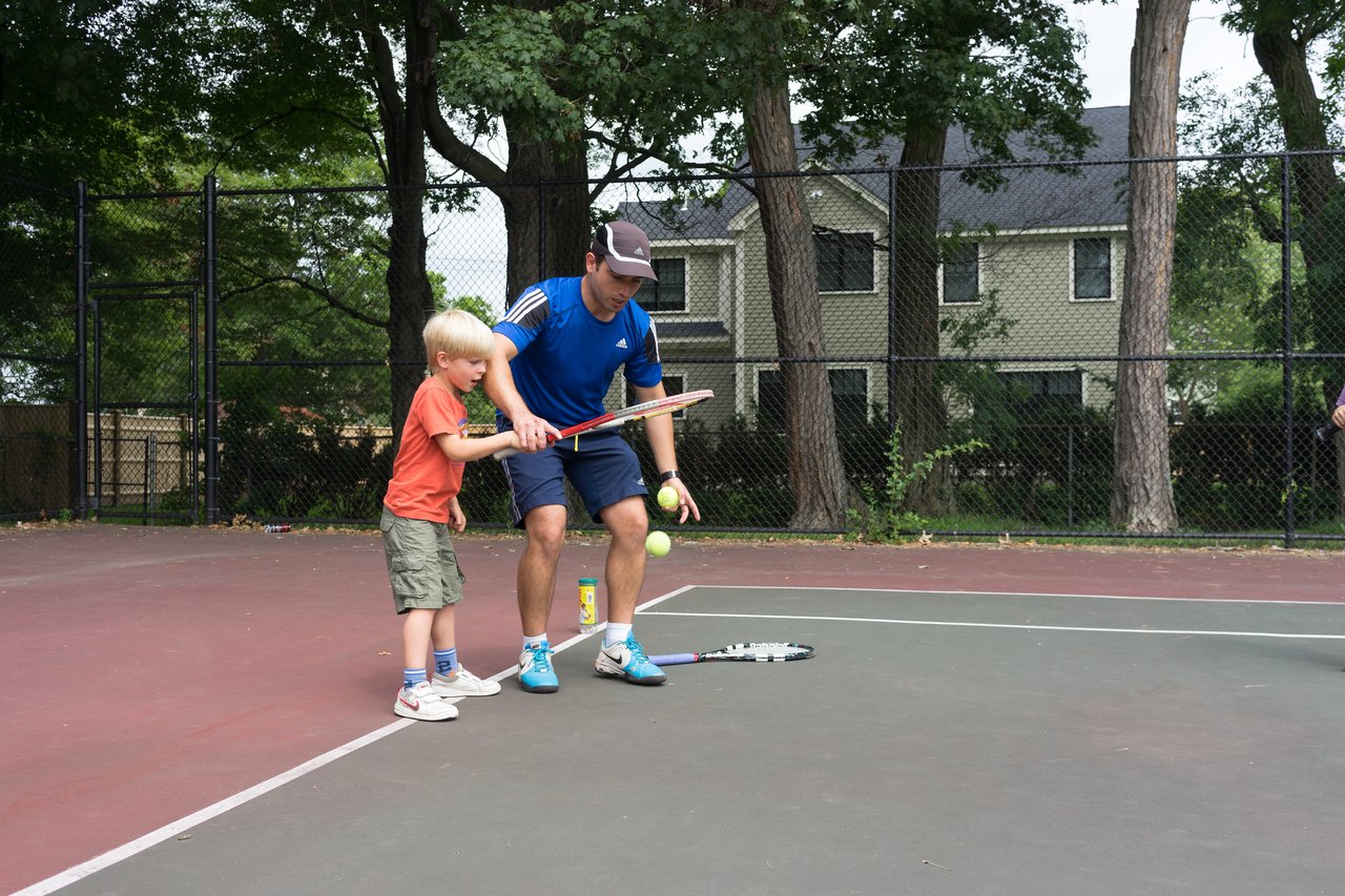 A tennis coach helps a young child practice holding a racket and hitting a ball on an outdoor court.