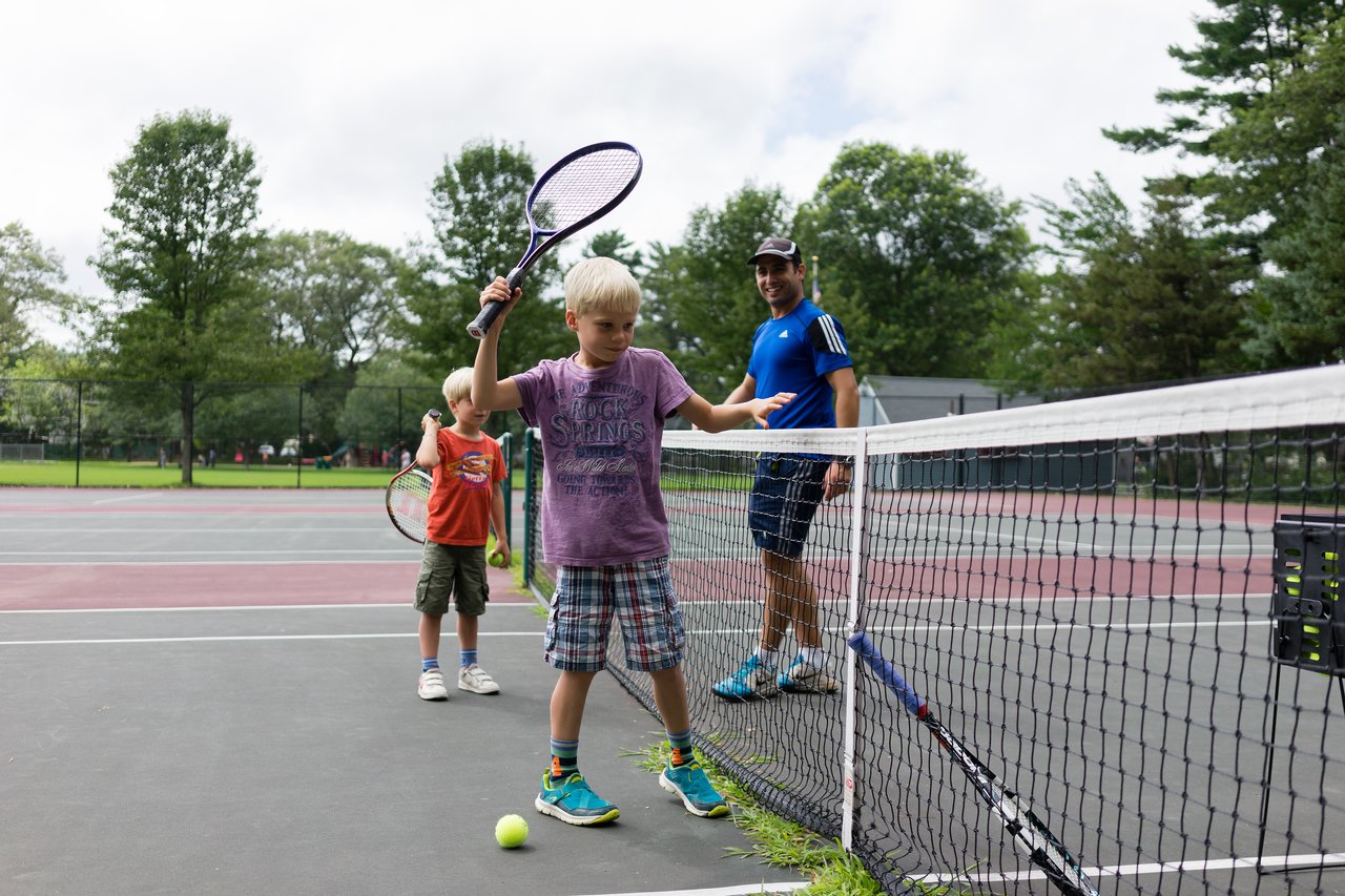 A young boy steps over a tennis net while holding a racket, with a coach and another child nearby.