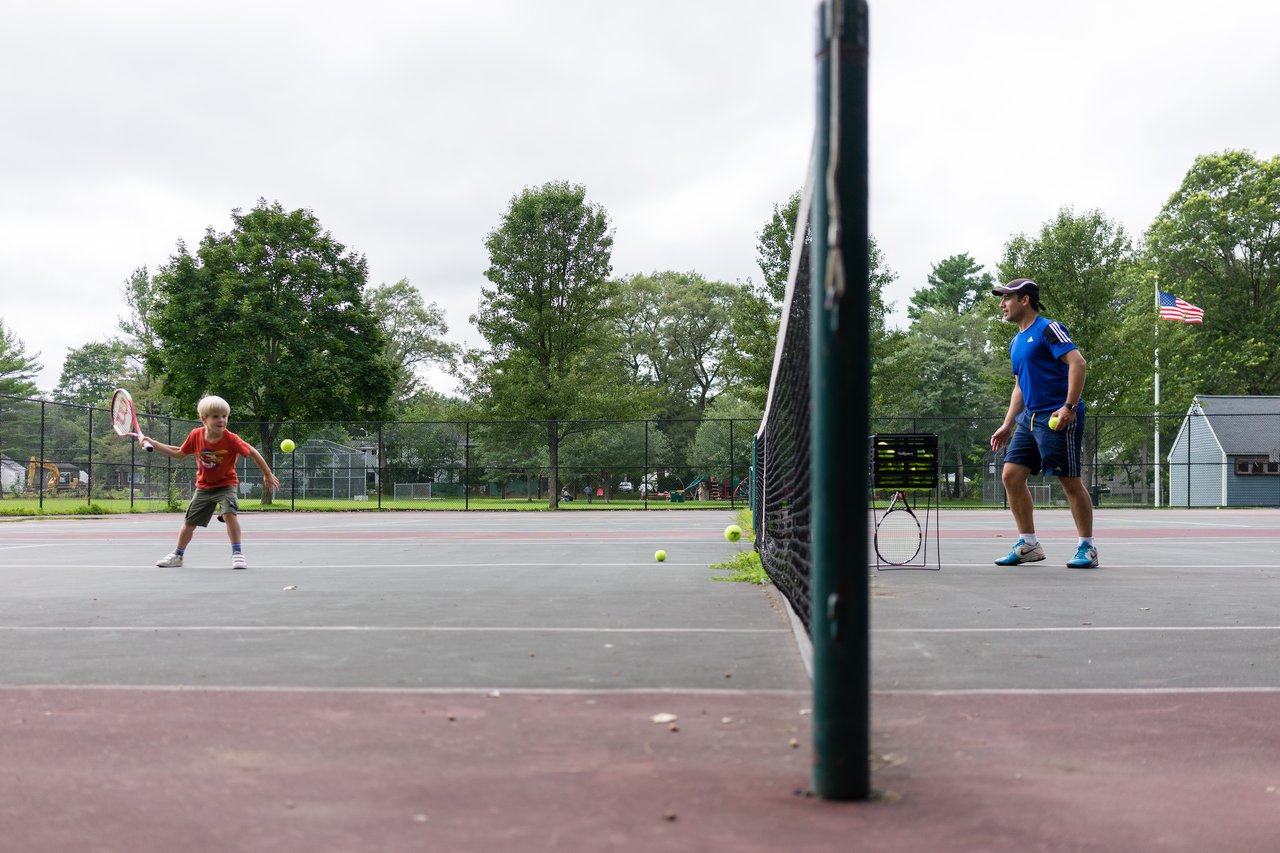 A young child practices hitting a tennis ball while an instructor watches from the other side of the net.