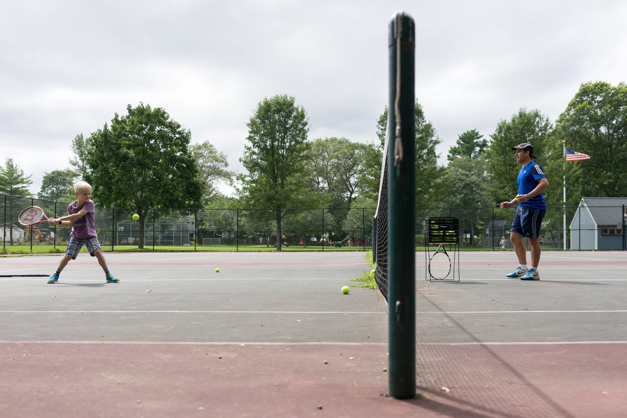 A child practices a backhand swing while a tennis coach observes on an outdoor court.