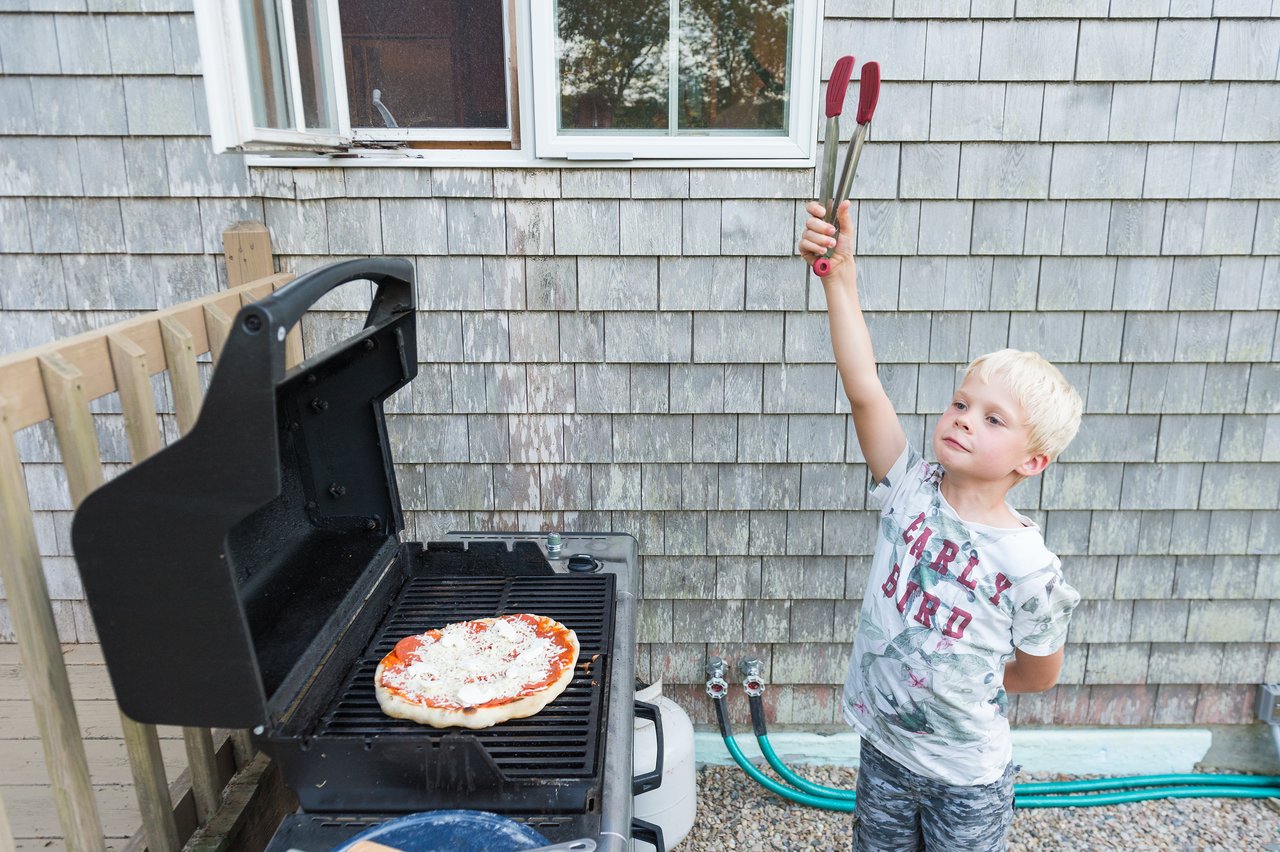 A young boy proudly holds up grilling tongs next to a barbecue with a pizza cooking on it.