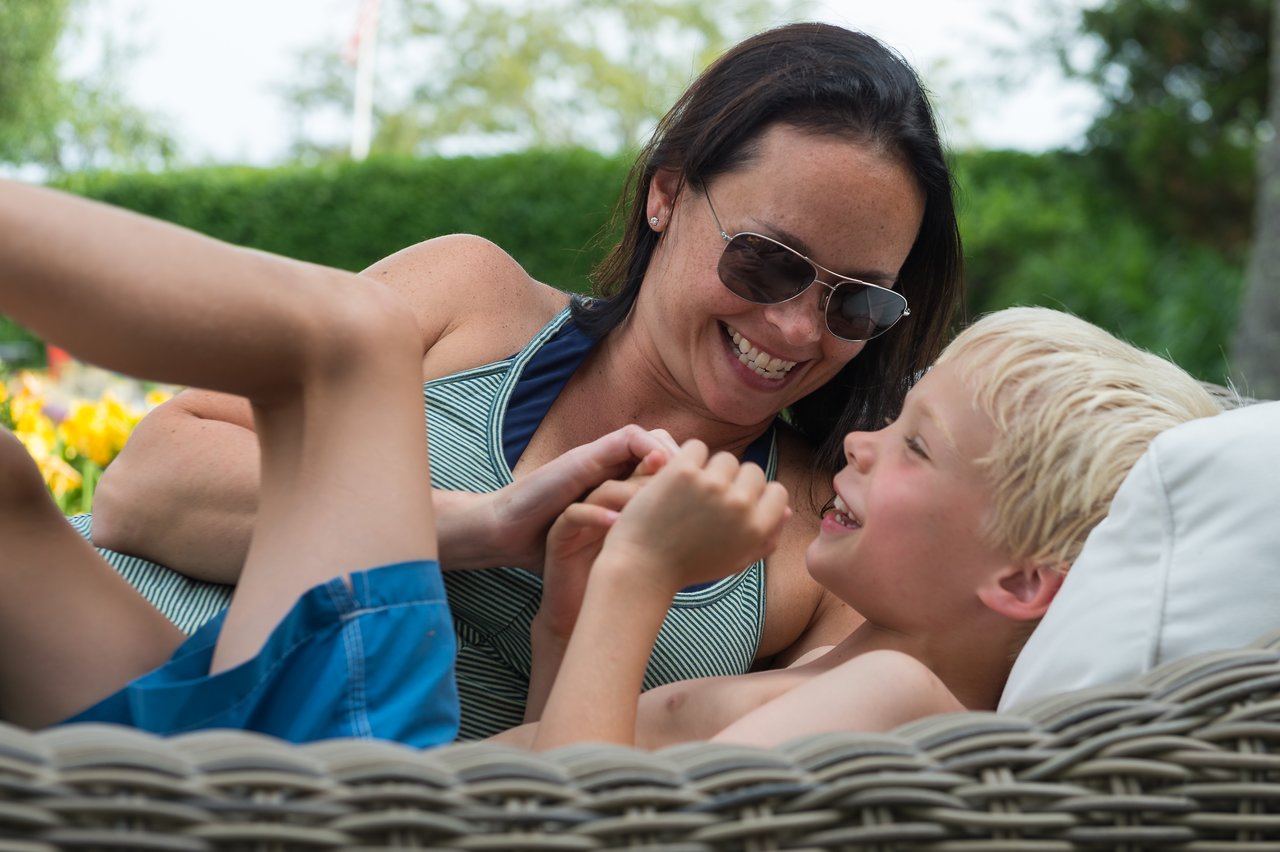 A woman and a young boy laugh together as she playfully tickles him on a wicker lounge chair.
