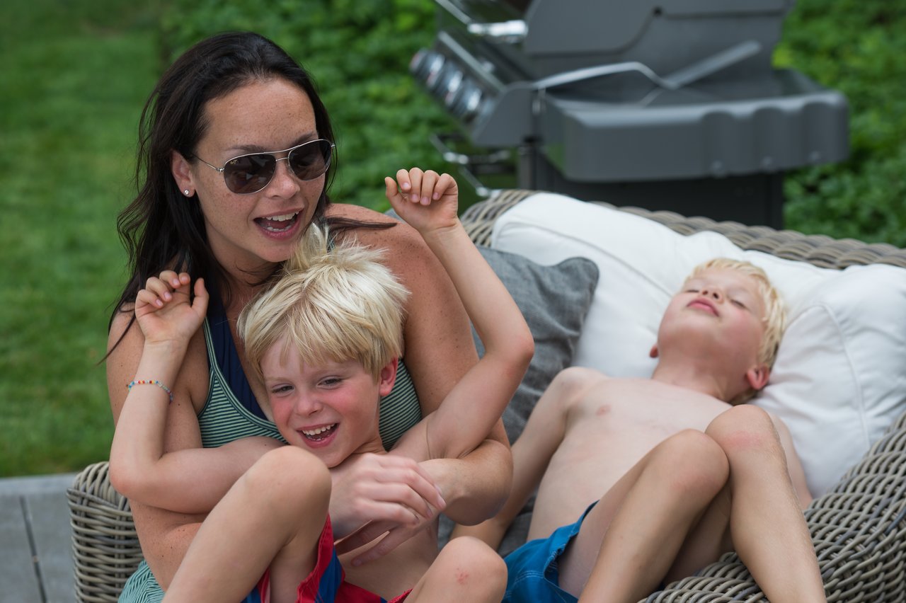 A woman playfully tickles a laughing child while another child relaxes on a wicker lounge chair.