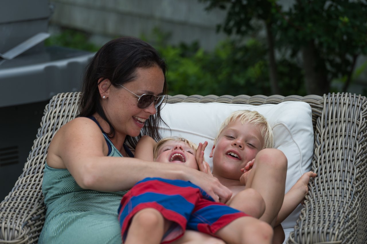 A woman playfully tickles two young boys on a lounge chair, making them laugh.