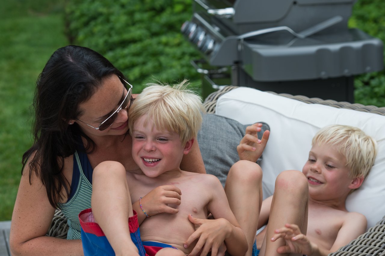 A woman playfully tickles a laughing child while another child watches and smiles on an outdoor couch.