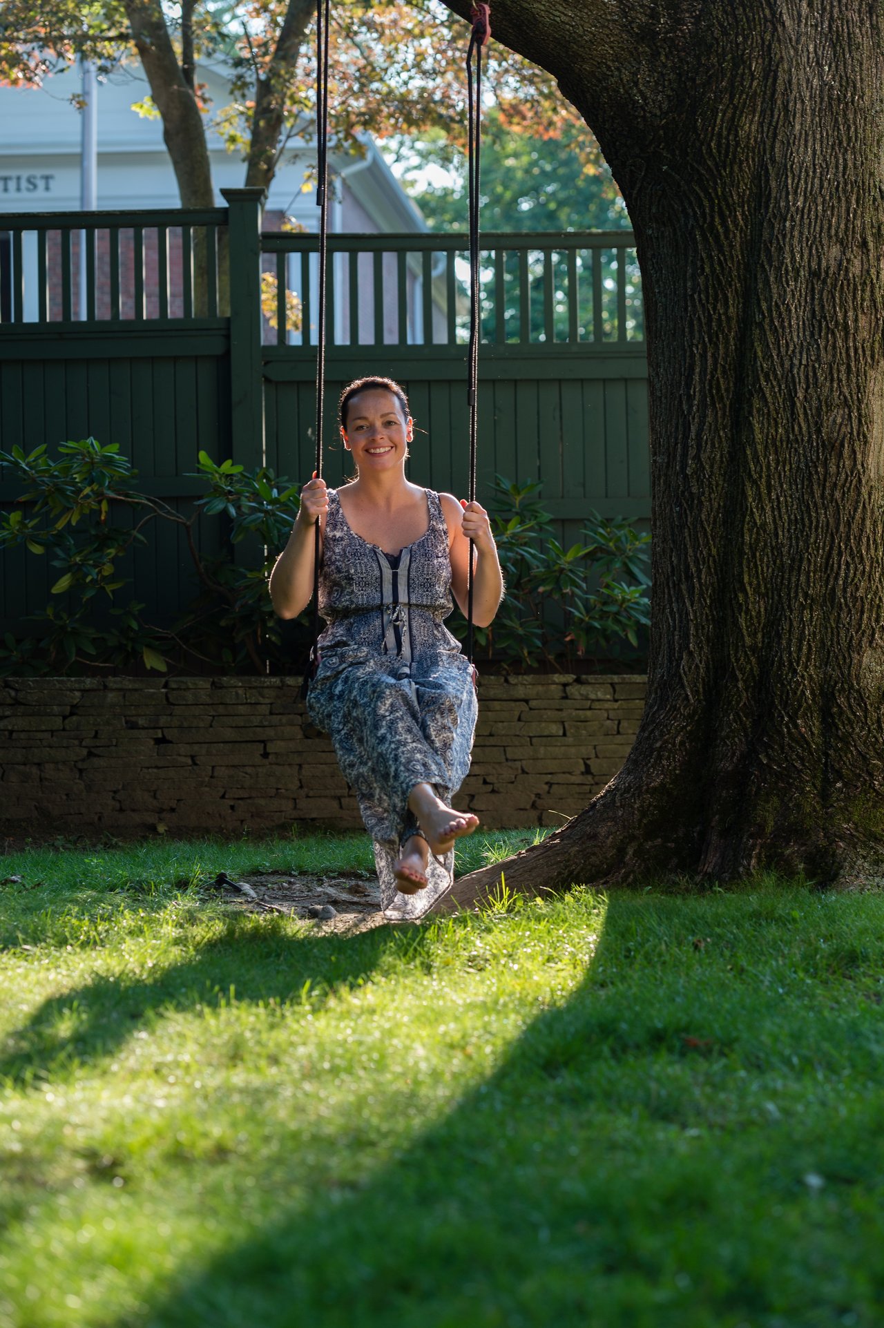 A woman sits on a tree swing, holding the ropes and smiling, with her bare feet slightly lifted.