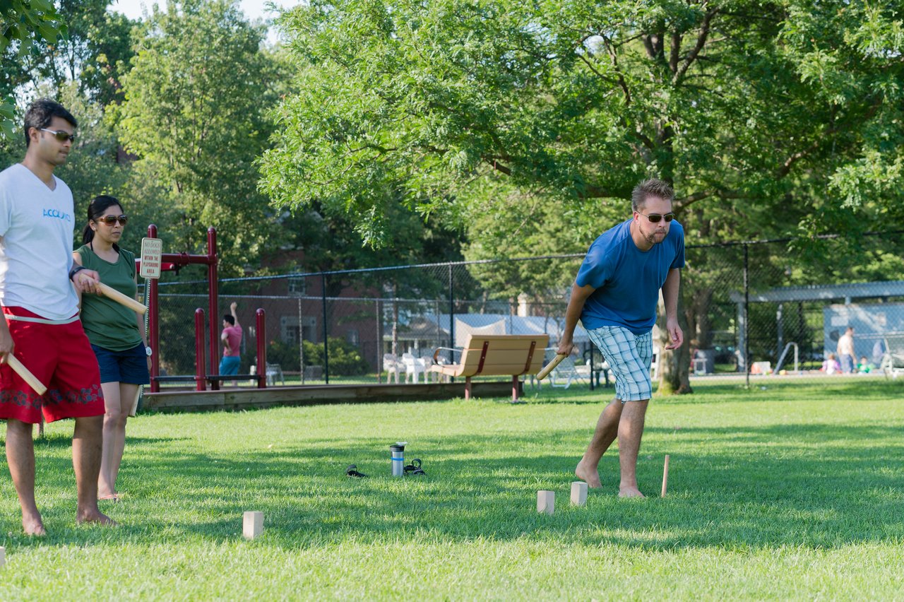 A man in a blue shirt throws a wooden stick while two others watch in a grassy park.