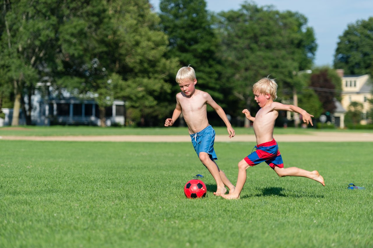 Two young boys play soccer on a grassy field, both running barefoot and focused on the red ball.