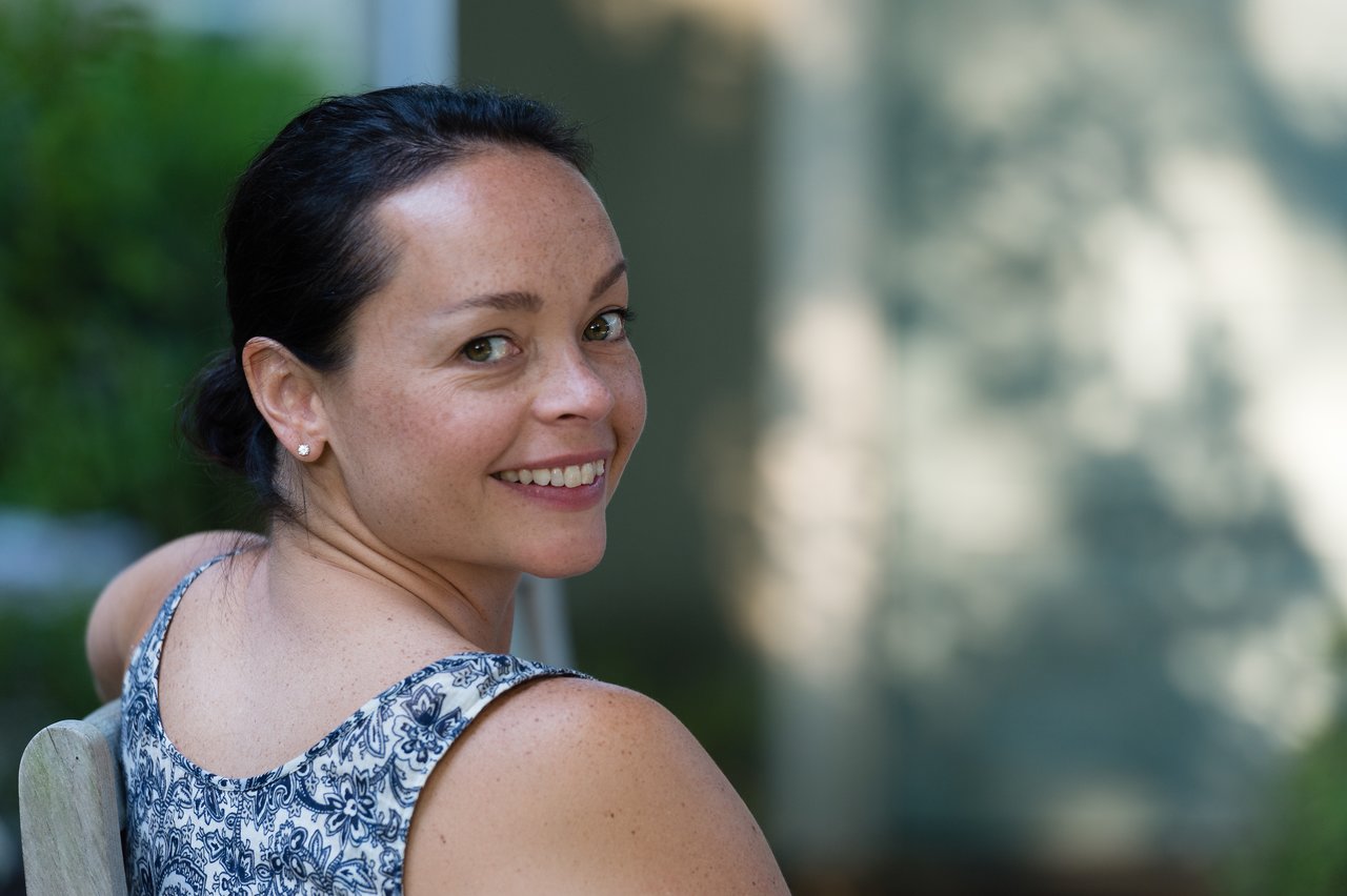 A woman with dark hair tied back smiles while turning to look at the camera, sitting outdoors.