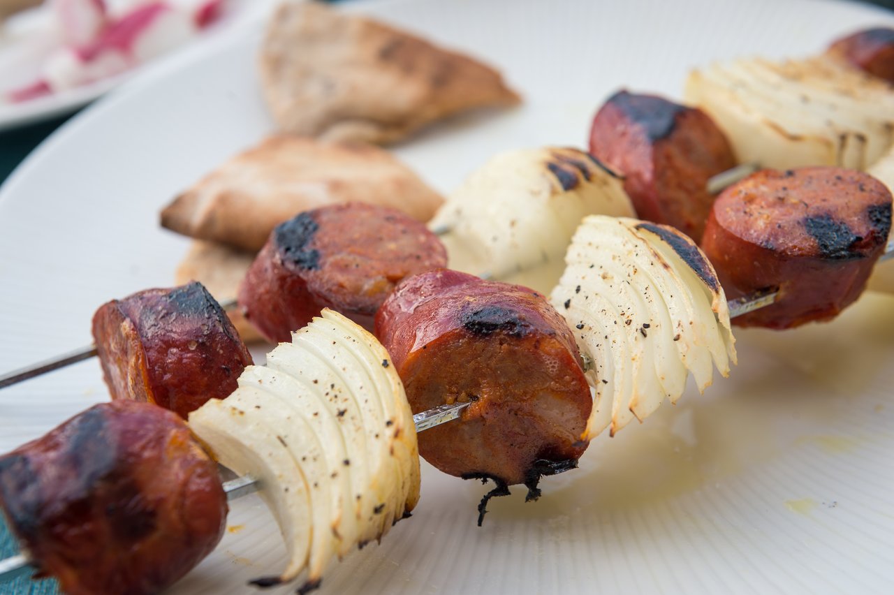 Grilled sausage and onion skewers on a white plate, with pita bread in the background.