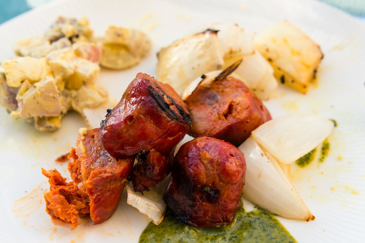 Grilled sausage and onions on a plate with potato salad and green sauce.