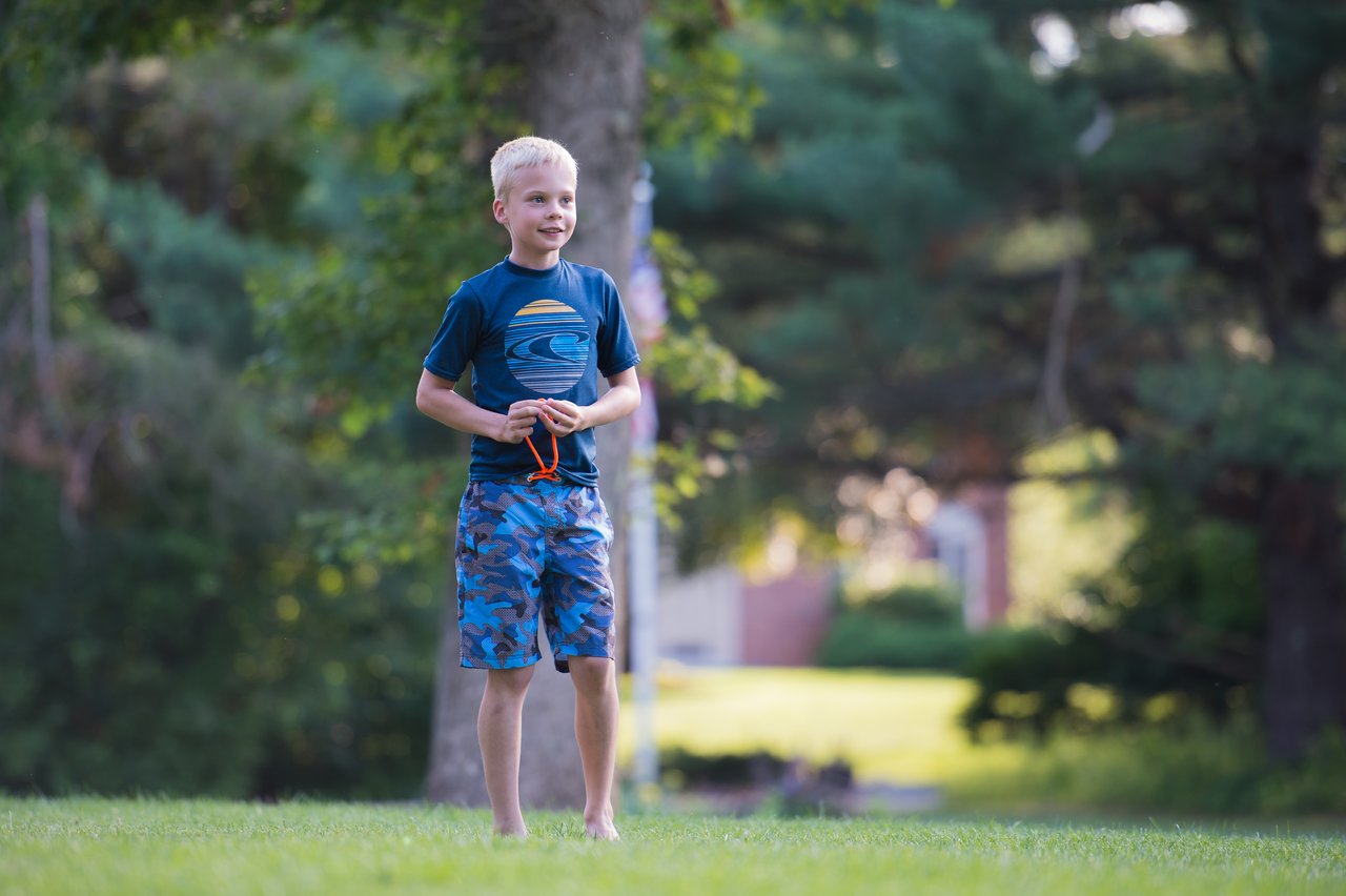 A young boy stands barefoot on the grass, smiling and looking ahead.