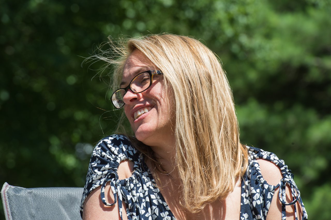 A woman with glasses and blonde hair smiles while sitting outdoors on a sunny day.