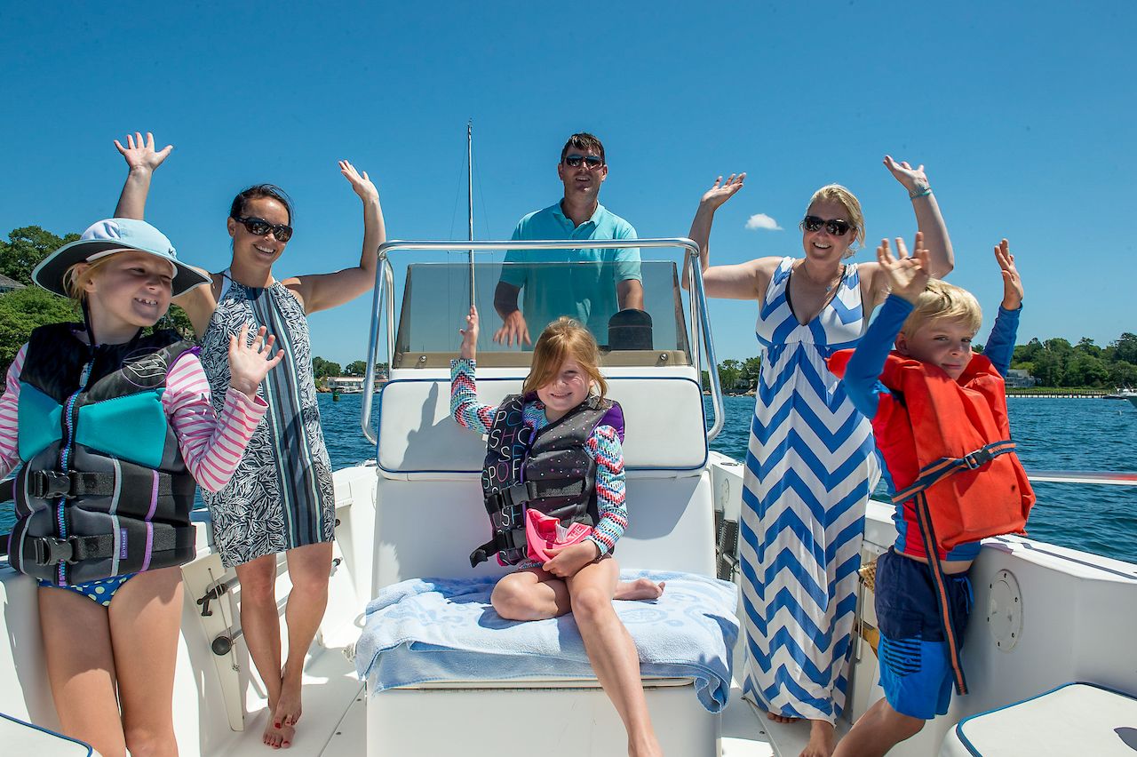 A group of adults and children wearing life jackets smile and raise their hands while boating on a sunny day.