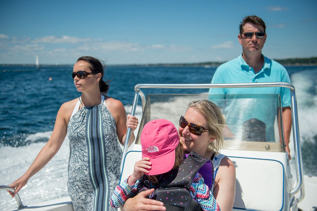 Four people ride on a motorboat; one woman hugs a child wearing a life jacket and pink hat.