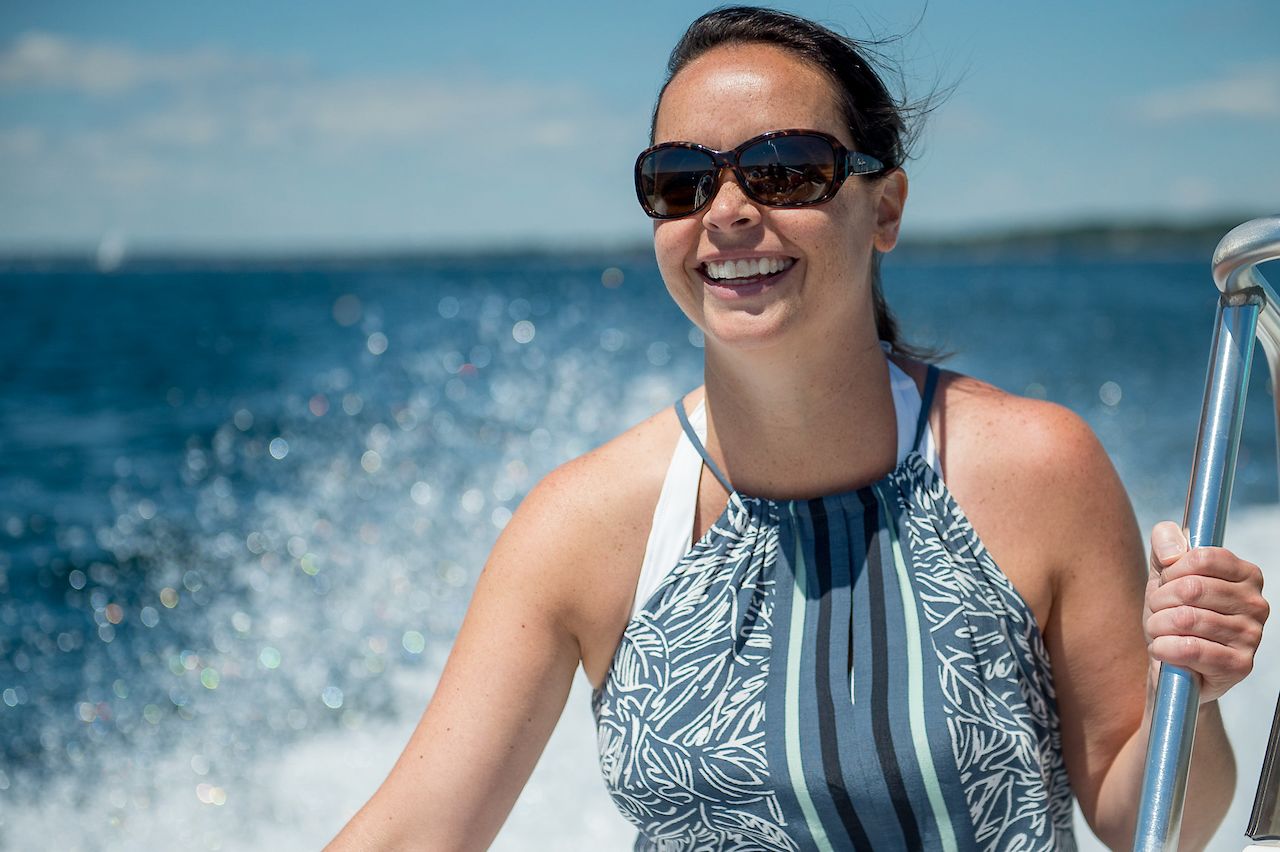 A smiling woman wearing sunglasses holds onto a metal railing while boating on the water.