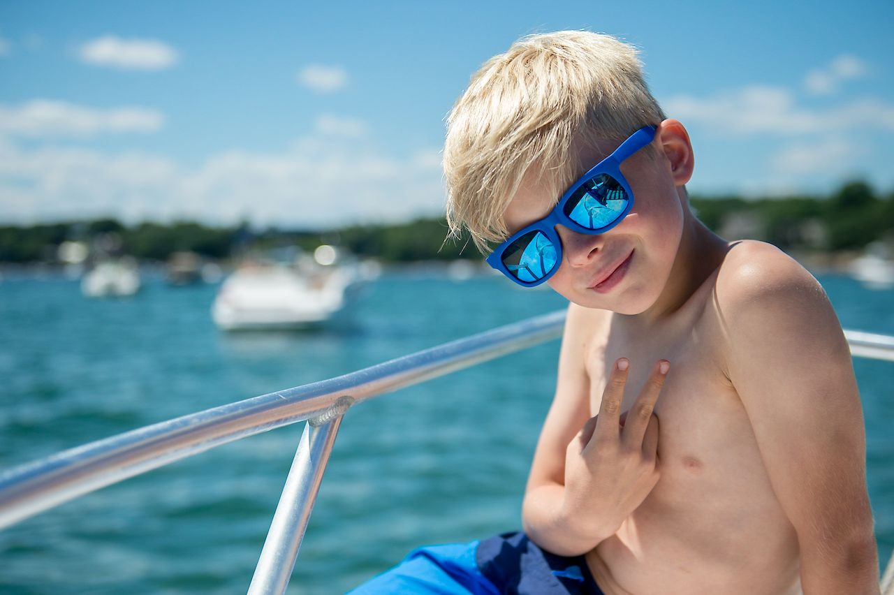 A young boy wearing blue sunglasses sits on a boat, smiling and making a peace sign with his fingers.