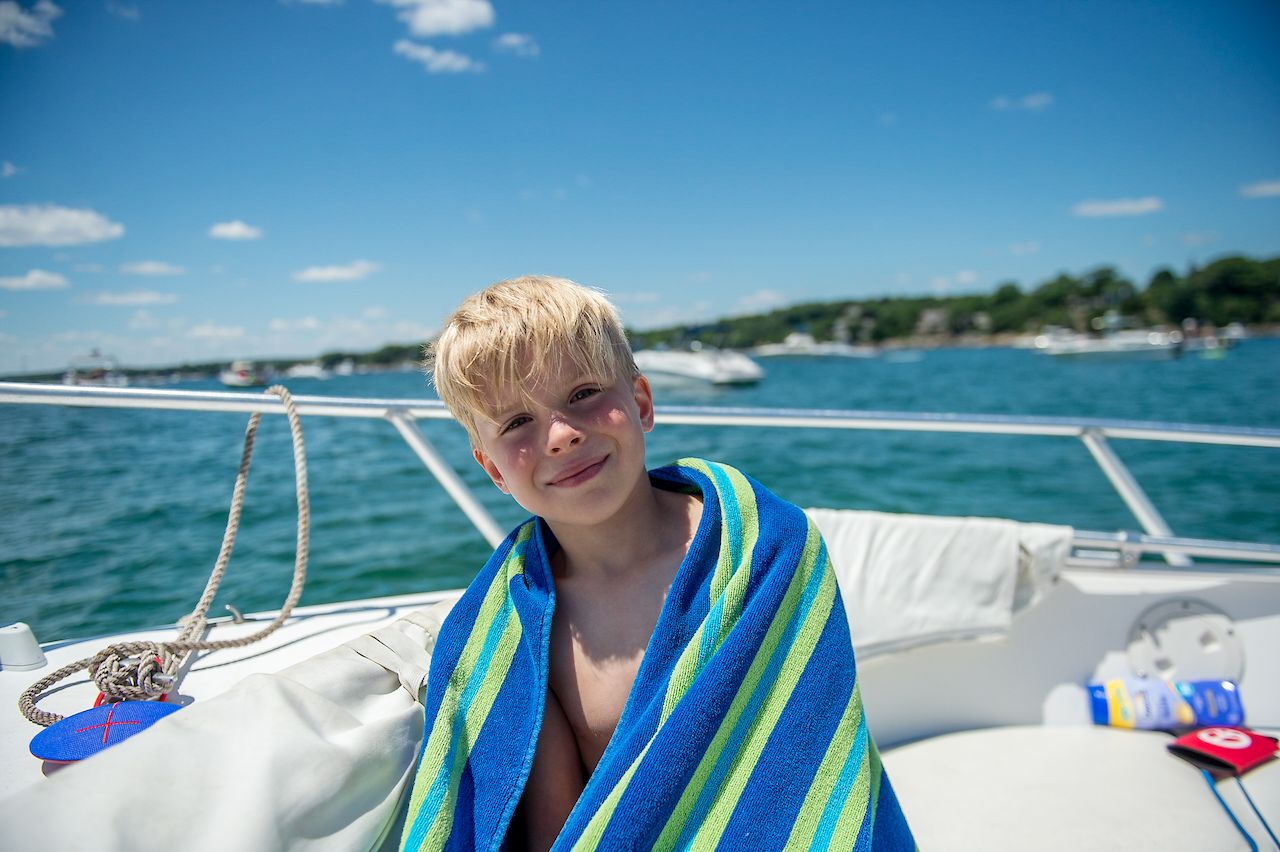 A young boy wrapped in a striped towel sits on a boat, smiling with the ocean and boats in the background.
