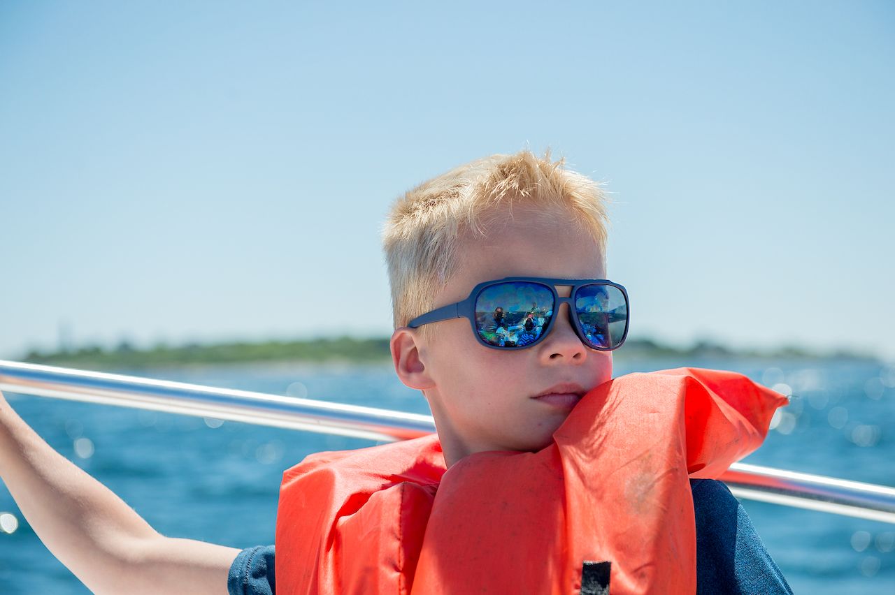 A child wearing sunglasses and a life jacket sits on a boat, looking ahead while holding onto the railing.