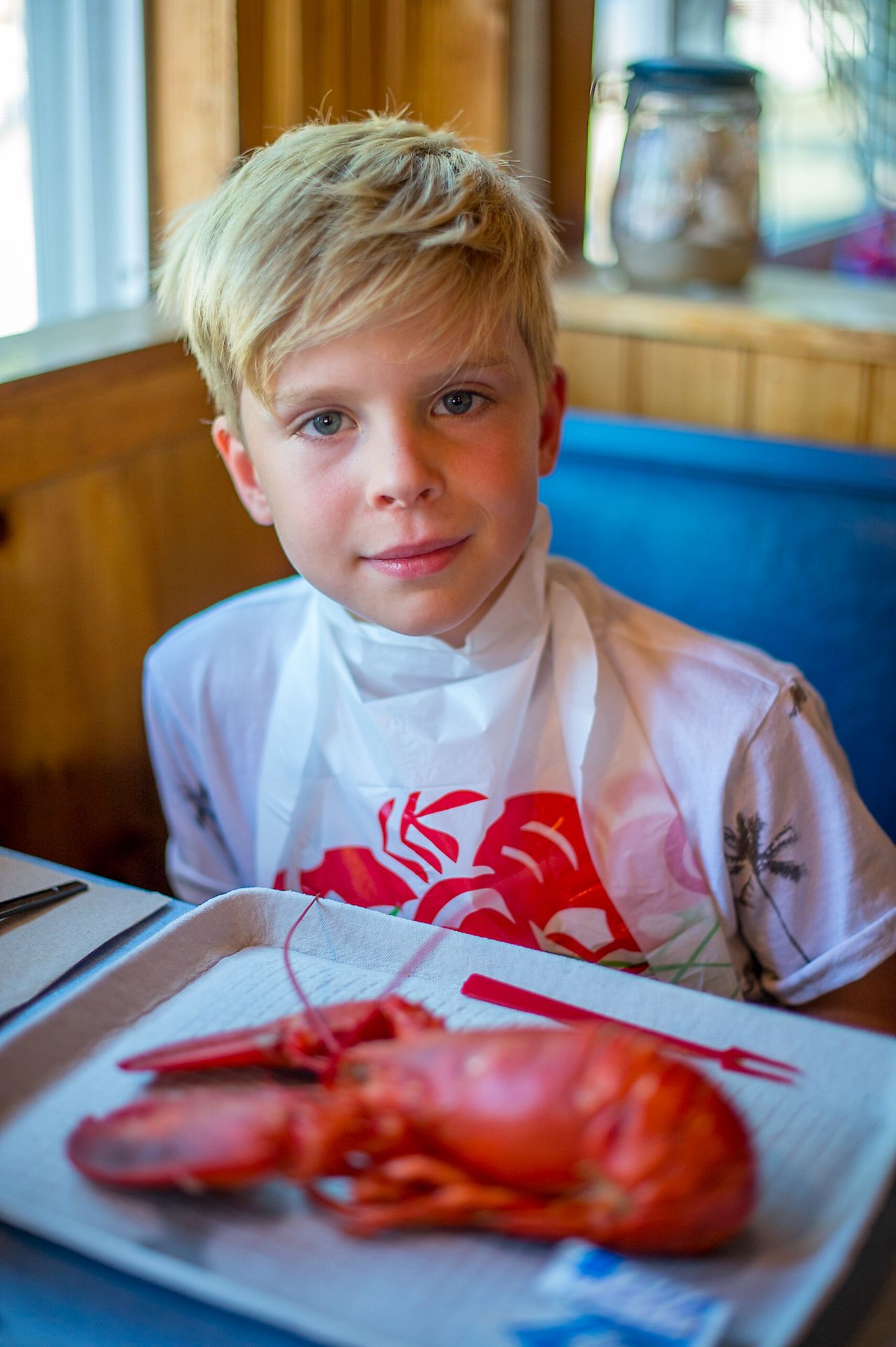A young boy wearing a lobster bib sits at a table with a cooked lobster on a tray.