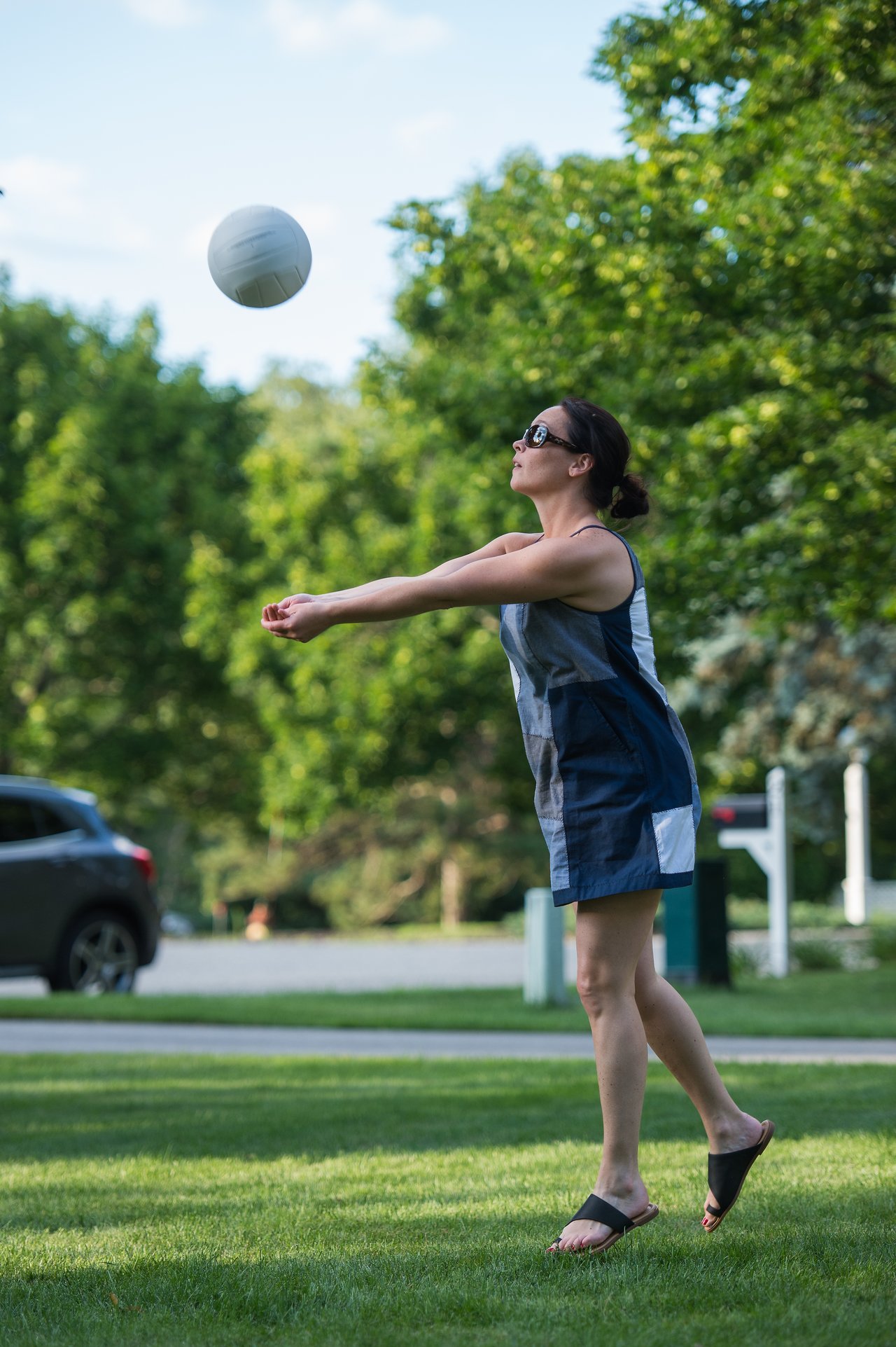 Vanessa bumps a volleyball with both arms while standing on grass, wearing sunglasses and a casual dress.