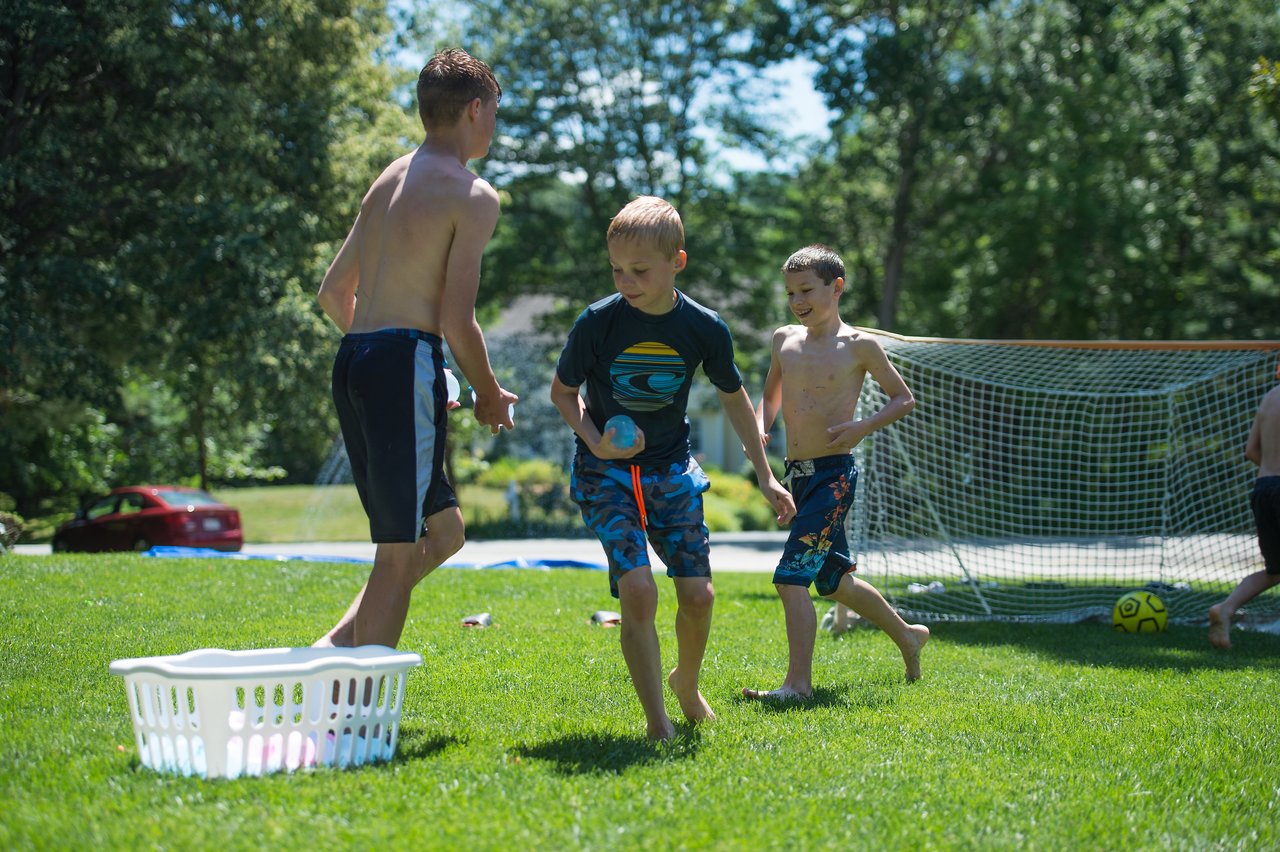 Three boys play with water balloons on a grassy field, running and laughing near a basket filled with balloons.