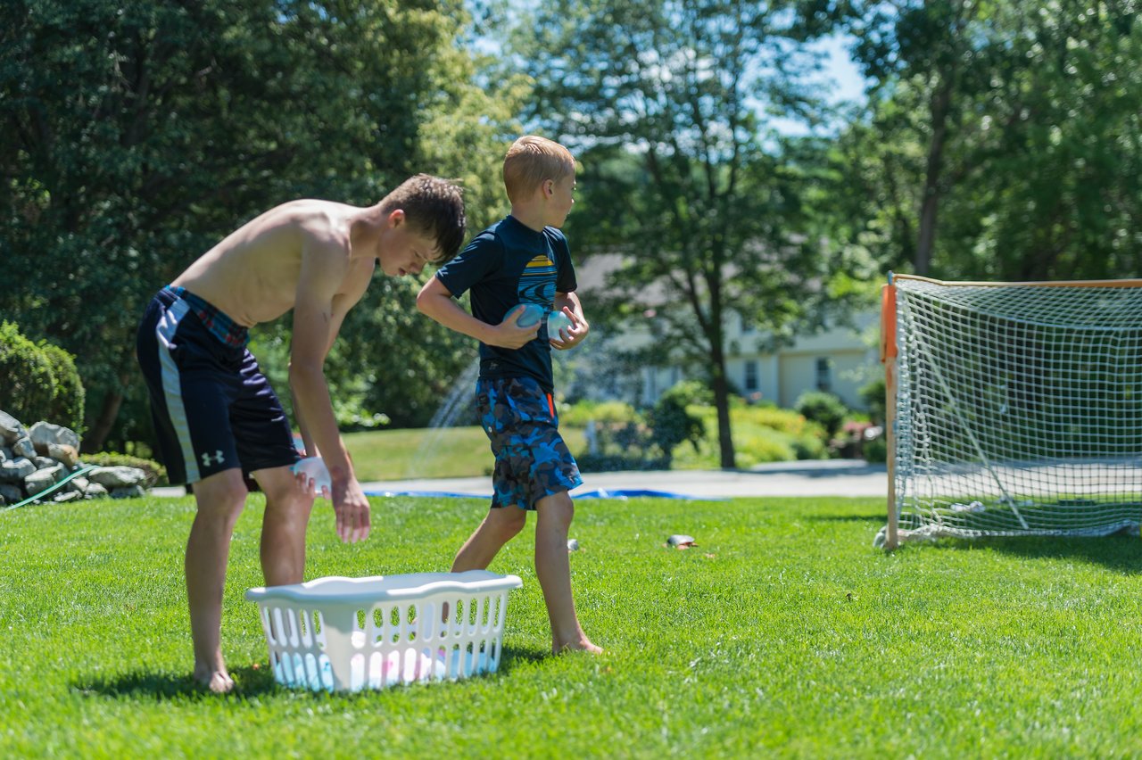 Two boys gather water balloons from a laundry basket on the grass, preparing for a water fight.