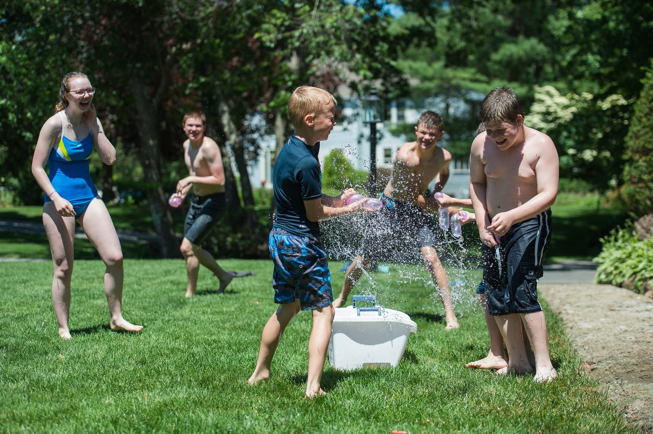 Children and teenagers play with water balloons on a sunny day, laughing as one bursts in mid-air.