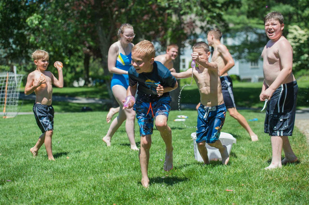 Children and teenagers play with water balloons on a sunny day, running and laughing on the grass.