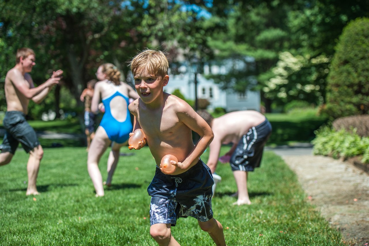 A boy runs across the grass holding two water balloons, while others play and splash in the background.