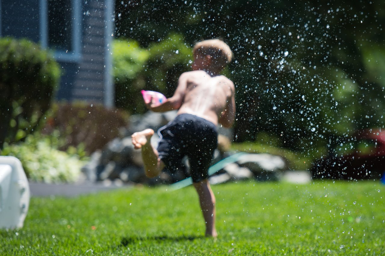 A child runs barefoot on the grass, holding a water balloon as droplets splash in the air.